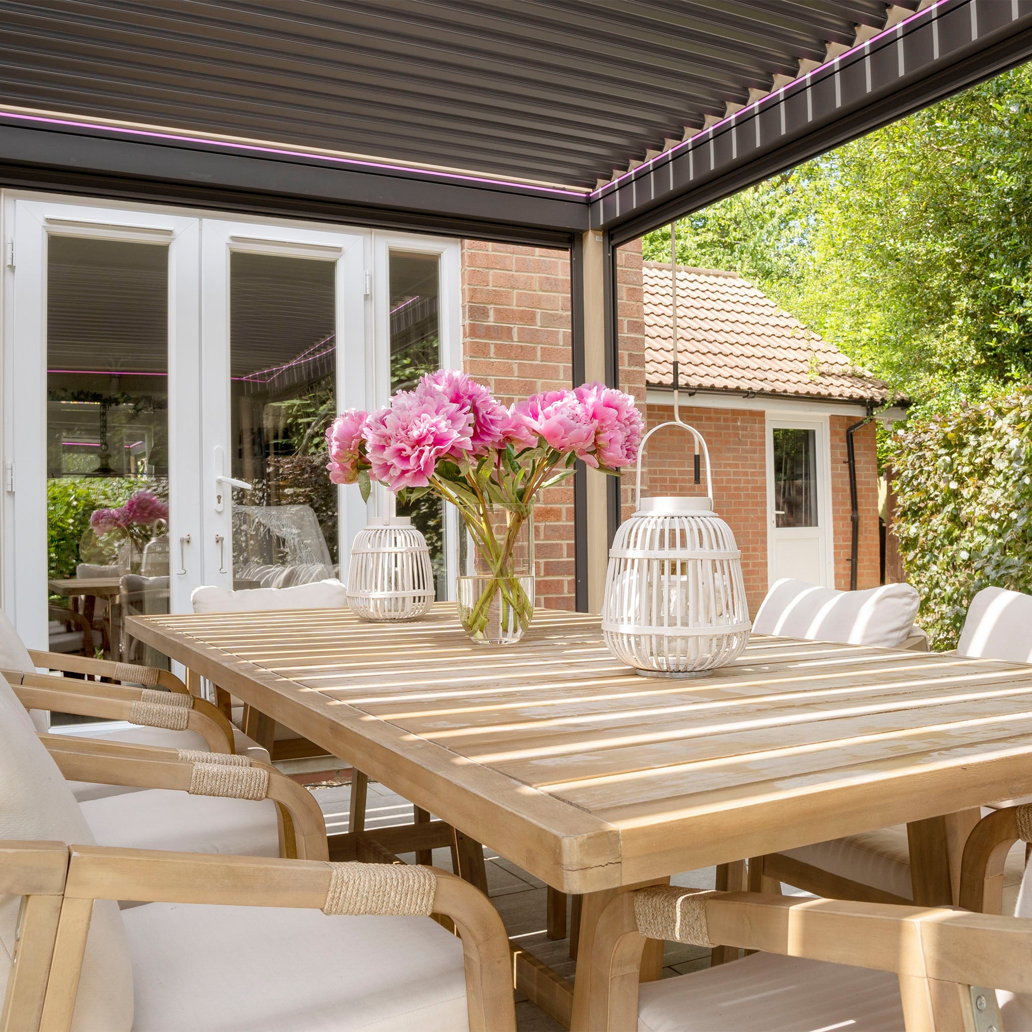 A patio table with pink flowers in a vase and white lanterns, surrounded by cushioned chairs under a PergoSTET Plus 3m x 4m Rectangular Pergola with adjustable louvres and LED lighting for ideal sun control.