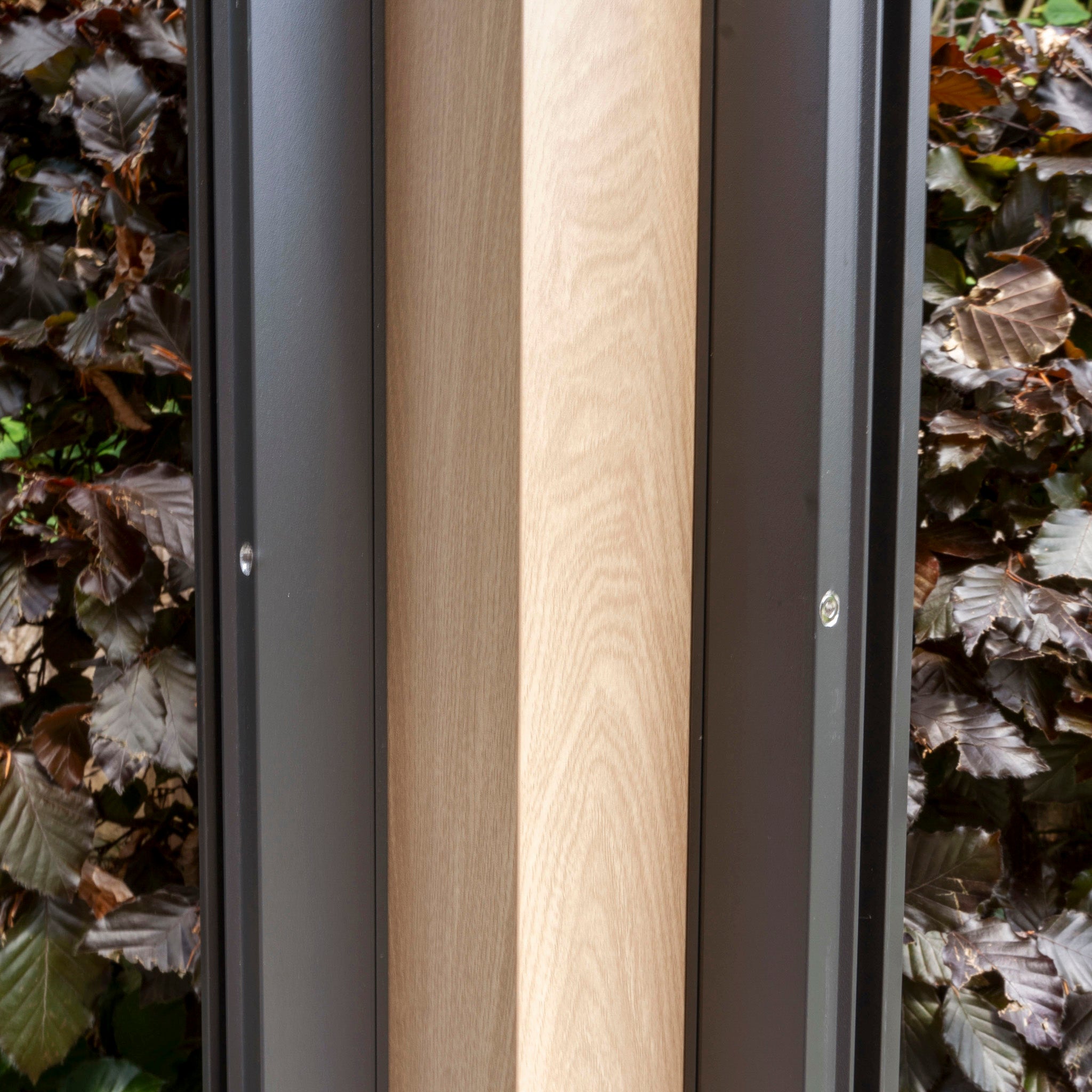 Close-up of a wooden post framed by two metal panels from the PergoSTET Plus 3m x 4m Pergola with wood effect, reminiscent of modern pergolas, with leafy bushes in the background.