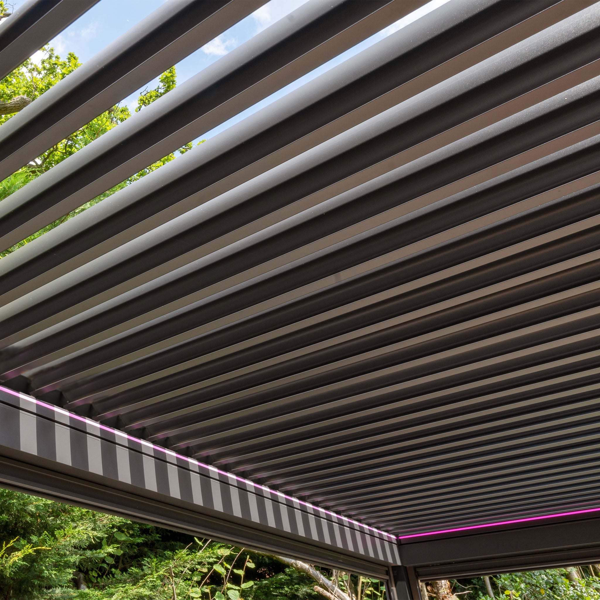 The PergoSTET Plus 3m x 3m Square Pergola in Grey features a sleek louvred roof, LED lighting, and includes three drop sides, beautifully set against a backdrop of trees.