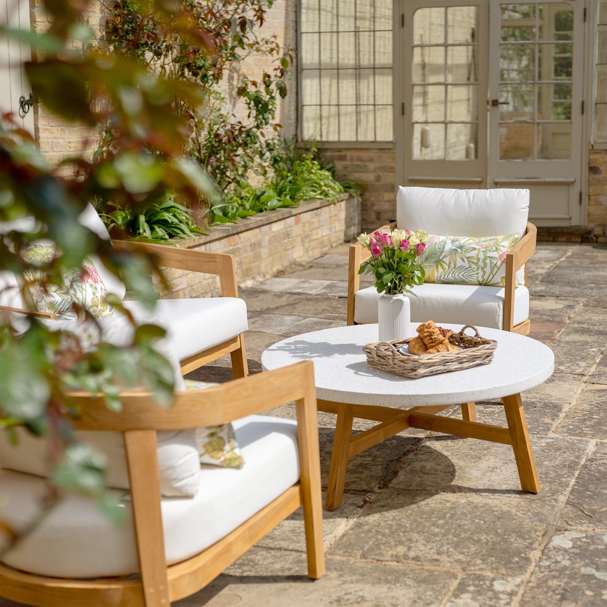 A cozy patio features a Yosemite 2 Seat Sofa Set in Glacier with premium teak wood chairs and water-resistant white polyester cushions, complemented by a round table with a basket and a potted plant from the Yosemite Collection.
