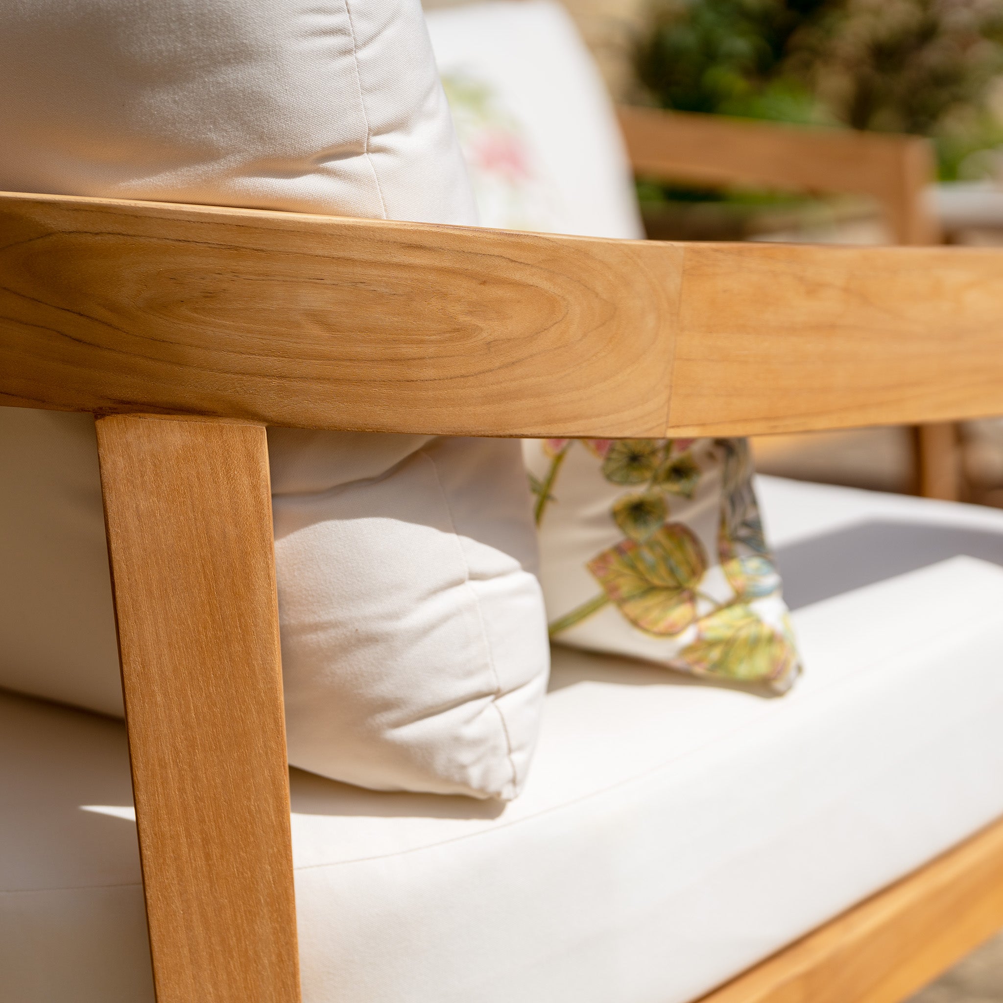 Close-up of a wooden outdoor 3-seat sofa from the Yosemite 3 Seat Sofa Set in Glacier, featuring white cushions and a floral-patterned pillow, set in a sunlit patio area. Crafted from sustainably sourced premium teak wood, it epitomizes elegant outdoor furniture.