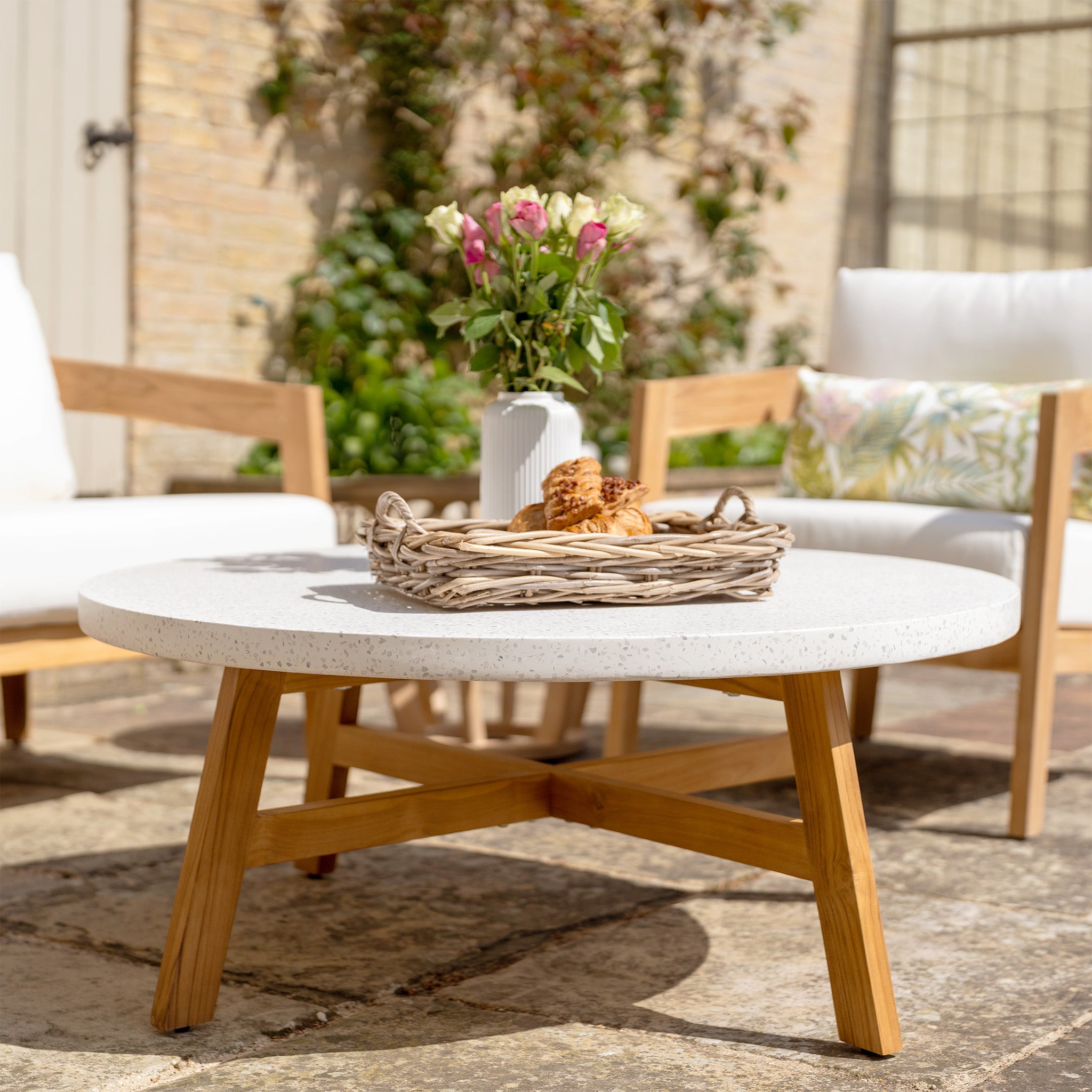 Outdoor patio with a stone table, wicker tray of pastries, wooden chairs with white cushions made from water-resistant polyester fabric, and flowers in a vase. This setup is part of the Yosemite 2 Seat Sofa Set In Glacier and features premium teak wood for an elegant touch.
