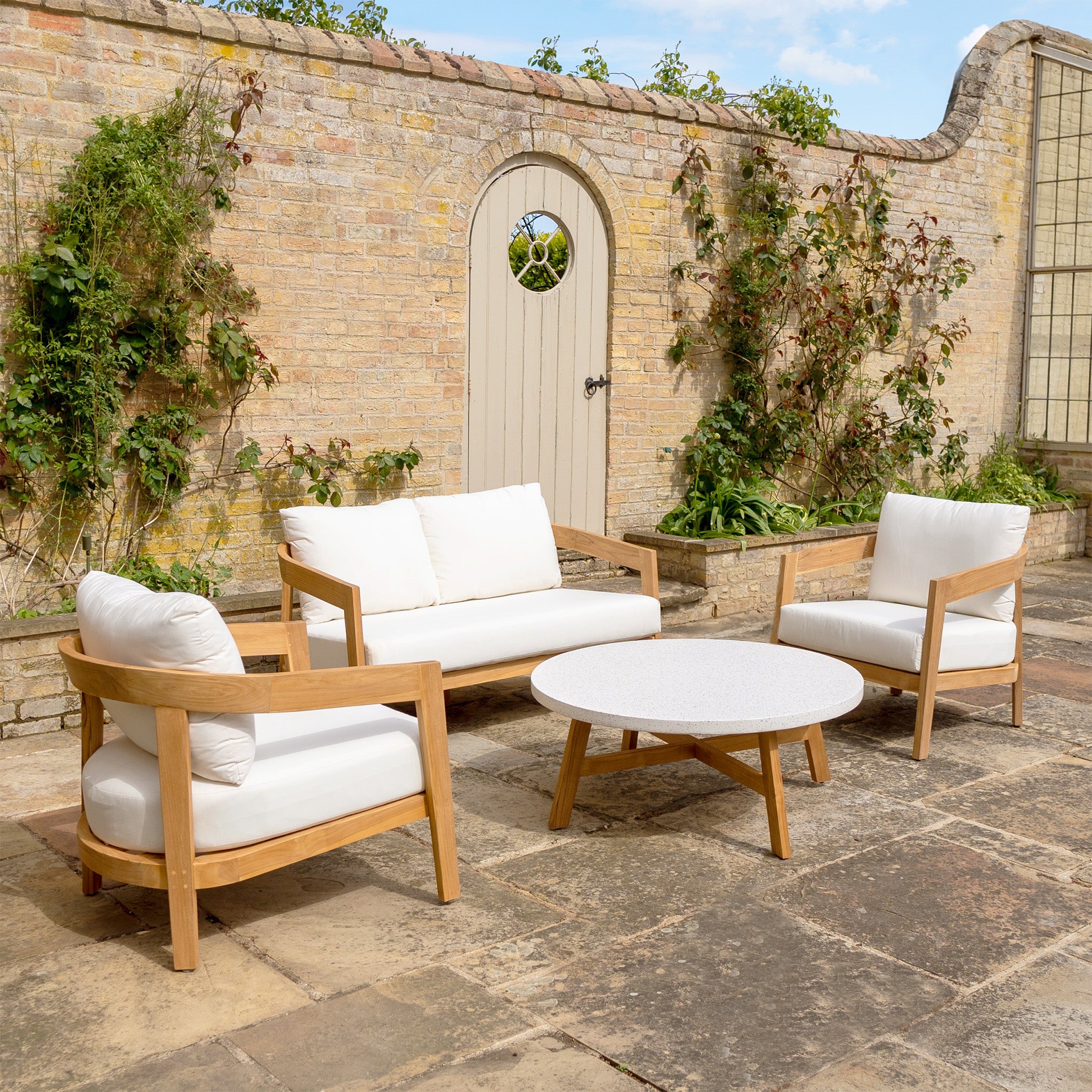 Outdoor patio with premium teak wood furniture, including a round table, two chairs, and the Yosemite 2 Seat Sofa Set in Glacier with water-resistant polyester fabric cushions, set against a brick wall.