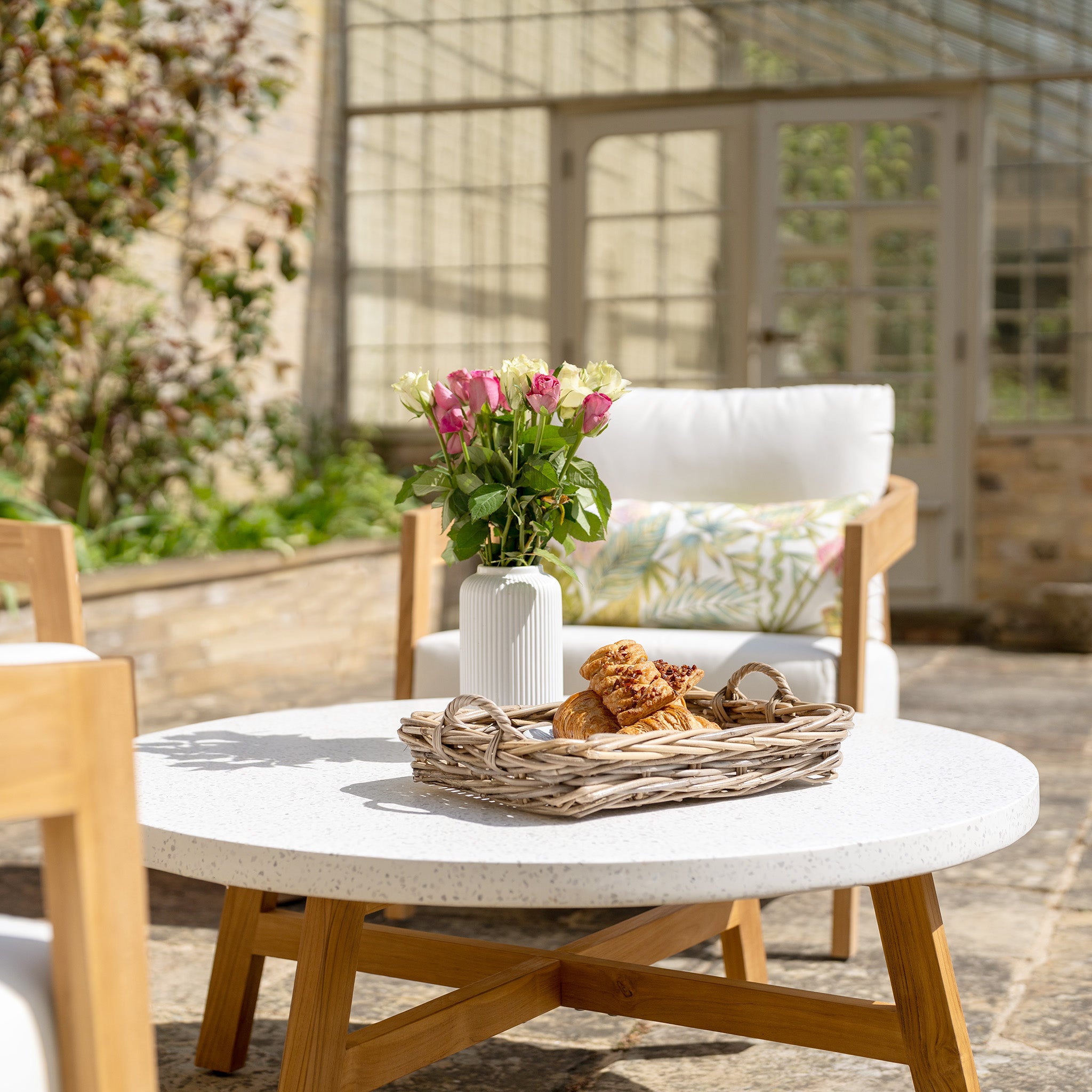 Outdoor patio featuring a table crafted from sustainably sourced premium teak wood holding a basket of pastries and a vase of roses, surrounded by the Yosemite 3 Seat Sofa Set in Glacier and lush greenery in the background.