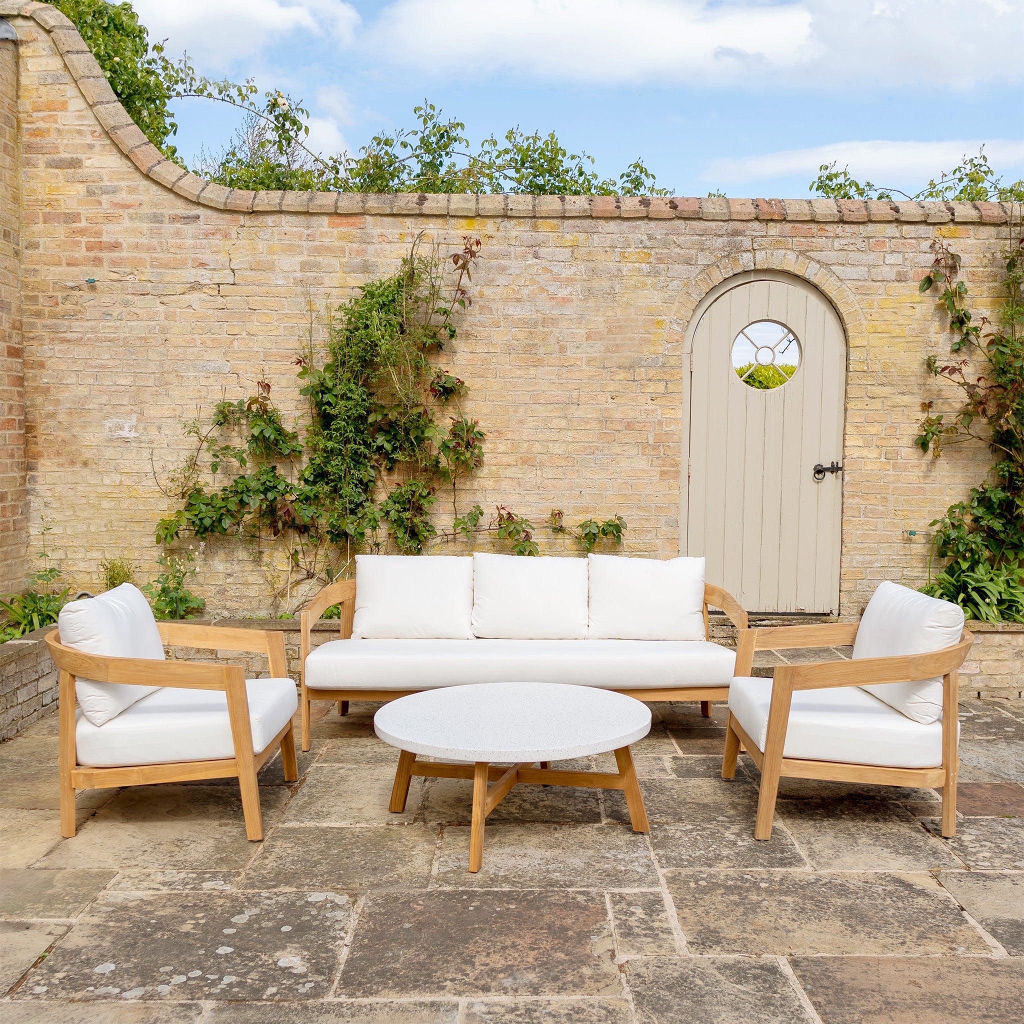 Introducing the Yosemite 3 Seat Sofa Set In Glacier: a cozy outdoor patio ensemble featuring a white sofa, two chairs crafted from sustainably sourced premium teak wood, and a round table against a brick wall with a wooden door.