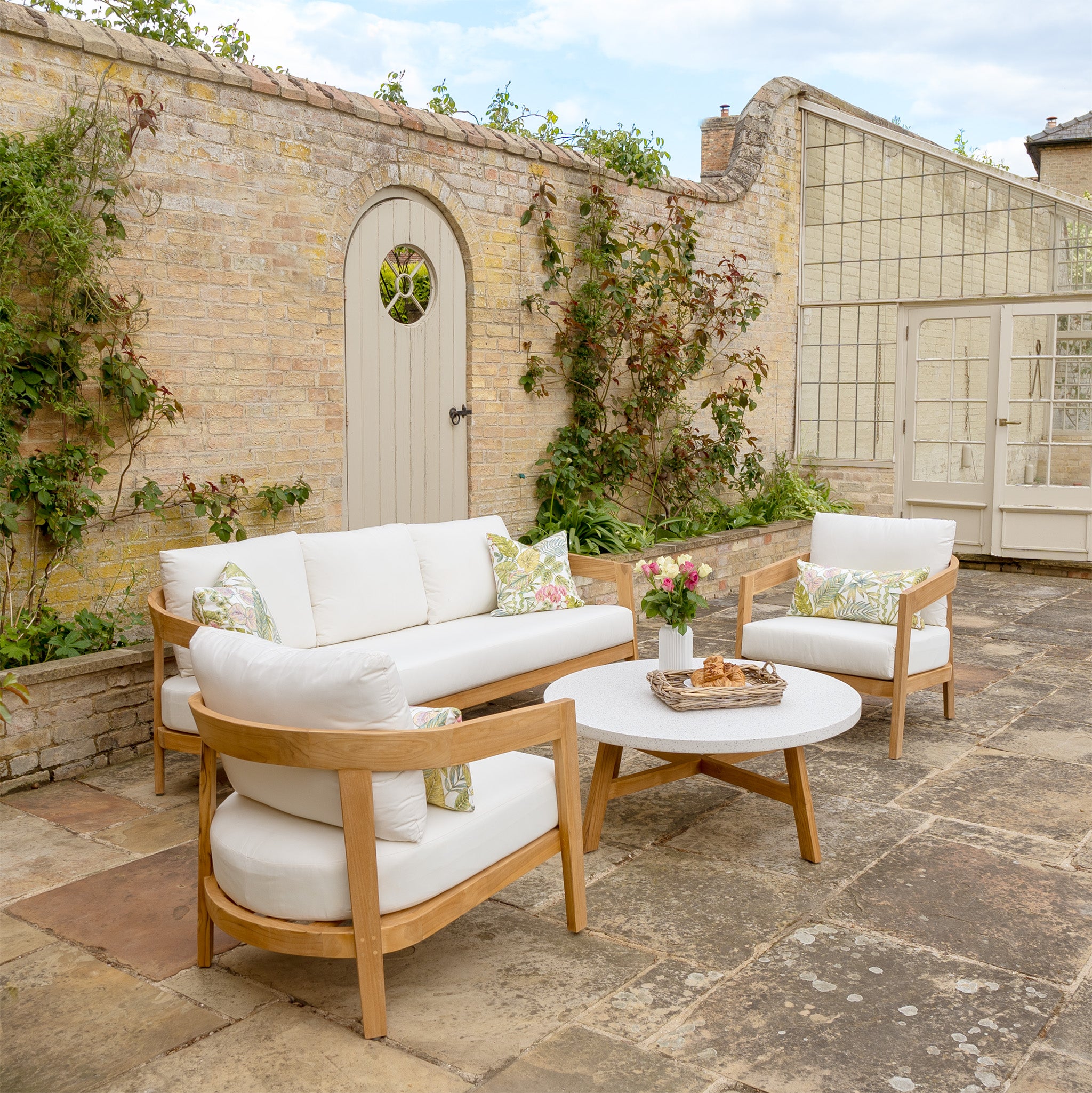 Cozy outdoor patio featuring the Yosemite 3 Seat Sofa Set In Glacier, with white cushioned wooden furniture, a round table, and a brick wall adorned with climbing plants. Each piece of this outdoor furniture set is crafted from sustainably sourced premium teak wood, adding both elegance and environmental responsibility.