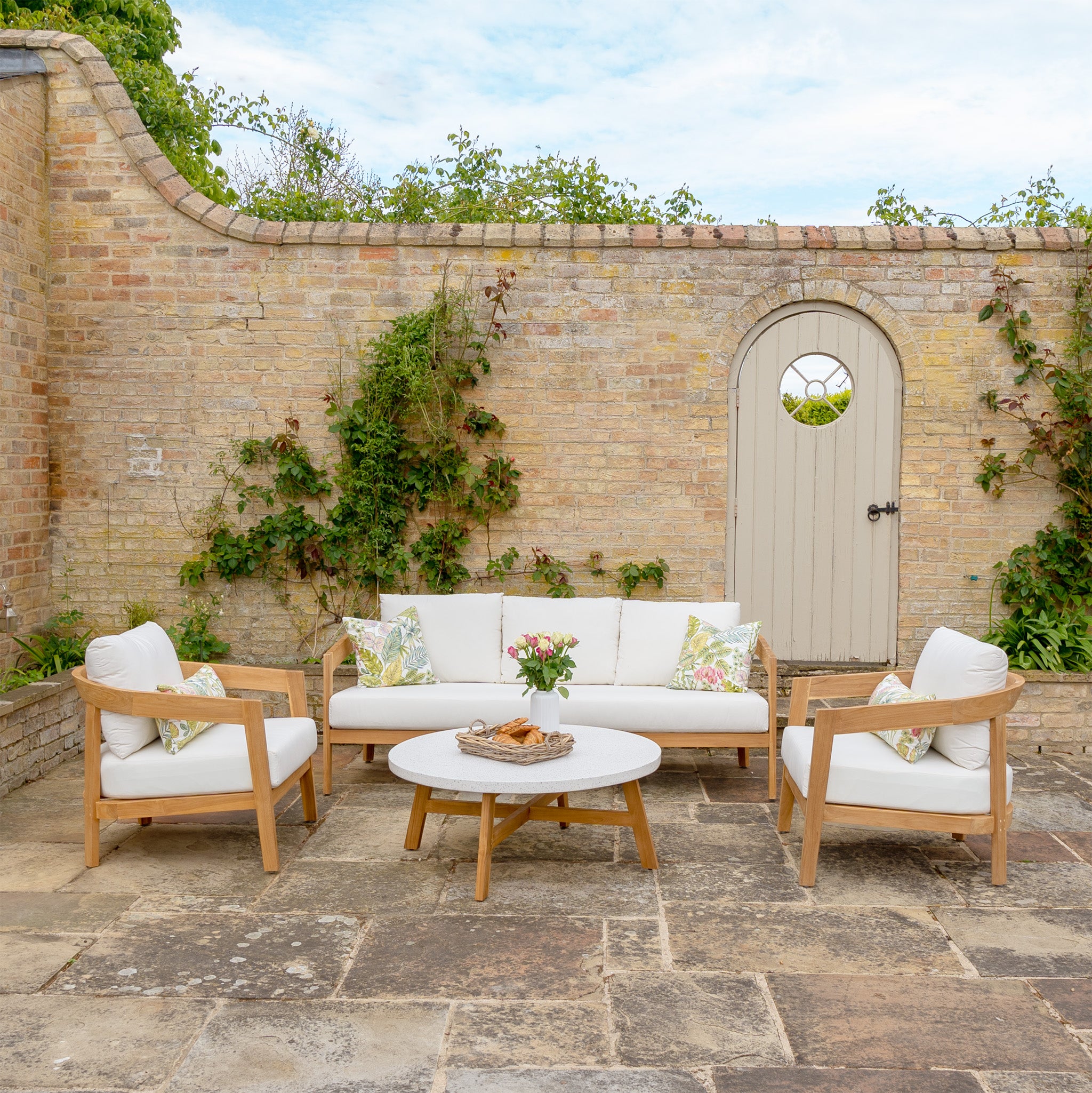 The Yosemite 3 Seat Sofa Set In Glacier from the Yosemite Collection, featuring light wood furniture crafted from sustainably sourced premium teak wood and adorned with white cushions, is set against a brick wall with greenery and an arched door.