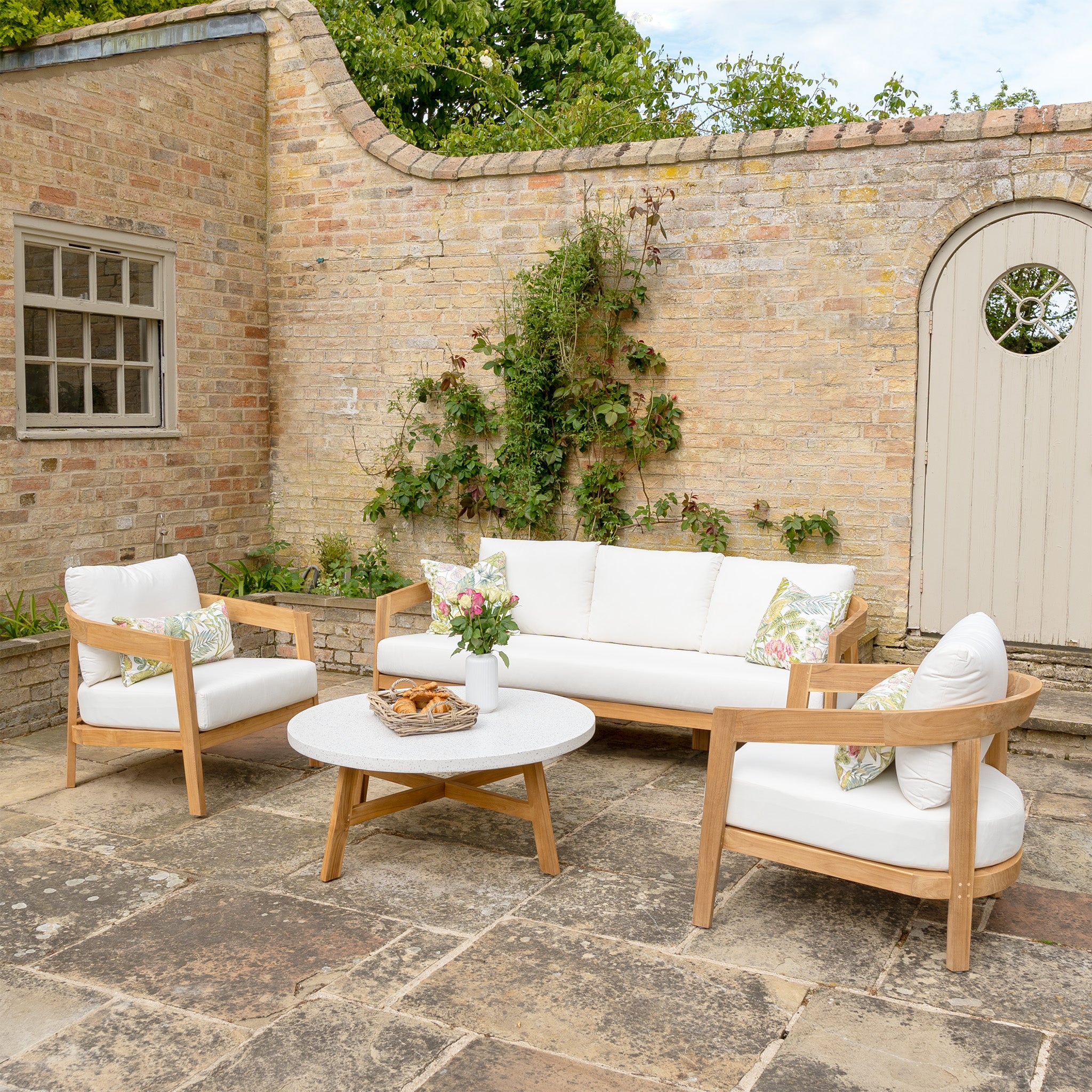 Introducing the Yosemite 3 Seat Sofa Set in Glacier, featuring white cushions, a wooden sofa, two chairs made from sustainably sourced premium teak wood, and a round table with snacks against a brick wall background.