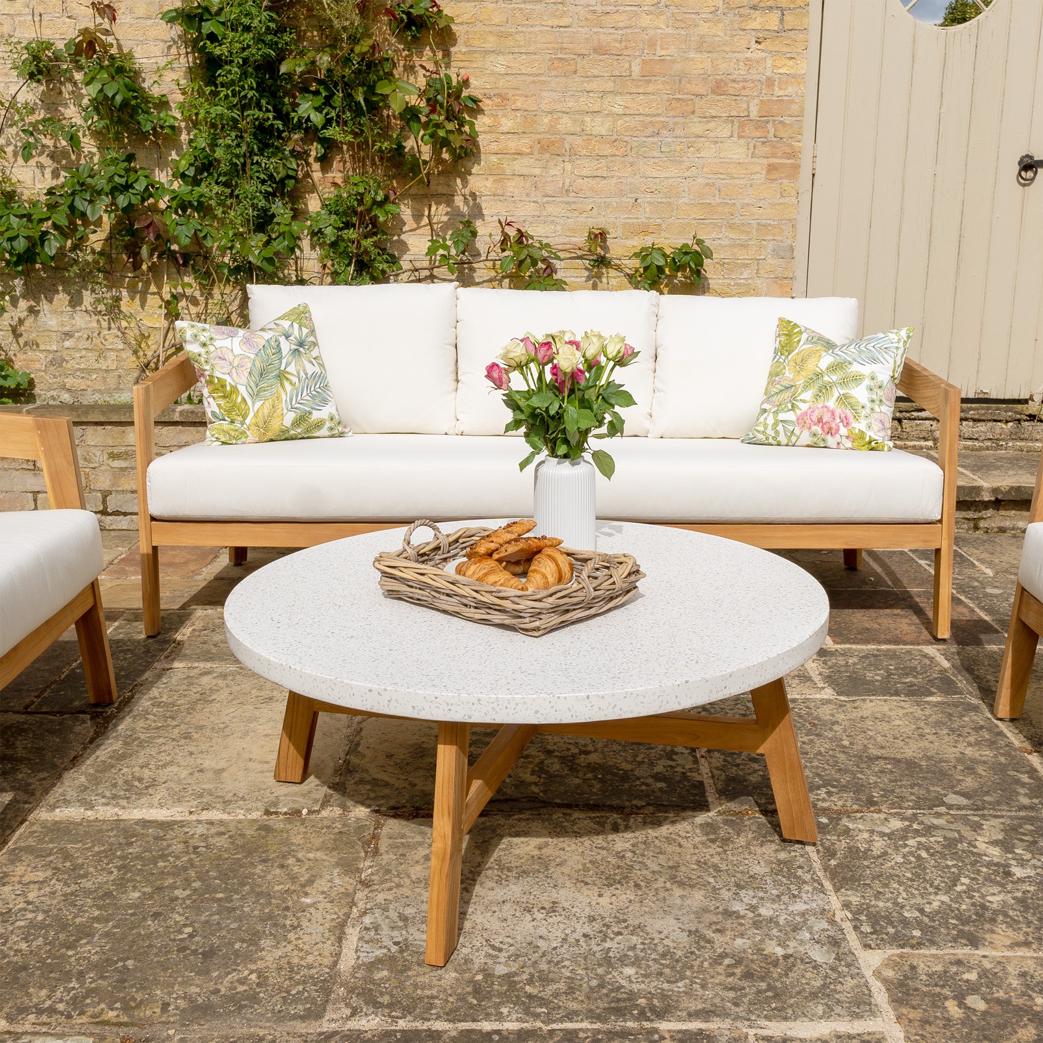 Outdoor patio featuring the Yosemite 3 Seat Sofa Set in Glacier, a round coffee table with pastries and flowers, set against a brick wall adorned with green plants. This elegant setup highlights our outdoor furniture crafted from sustainably sourced premium teak wood.
