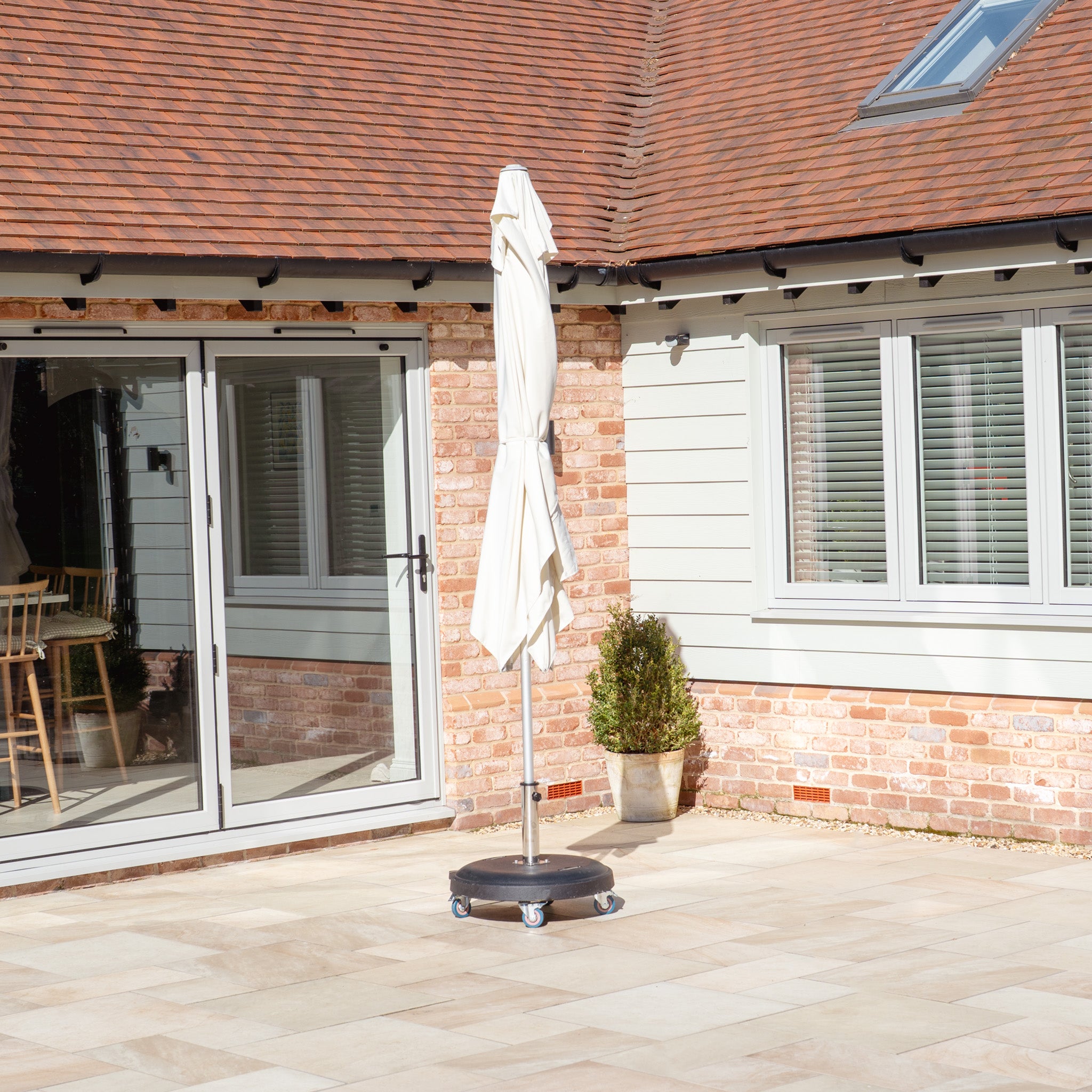 The Zelos 3m x 2m Rectangular Parasol with LED Lighting in Cream, on a wheeled base, stands closed by a house with large windows in a brick courtyard and offers UV protection when open.