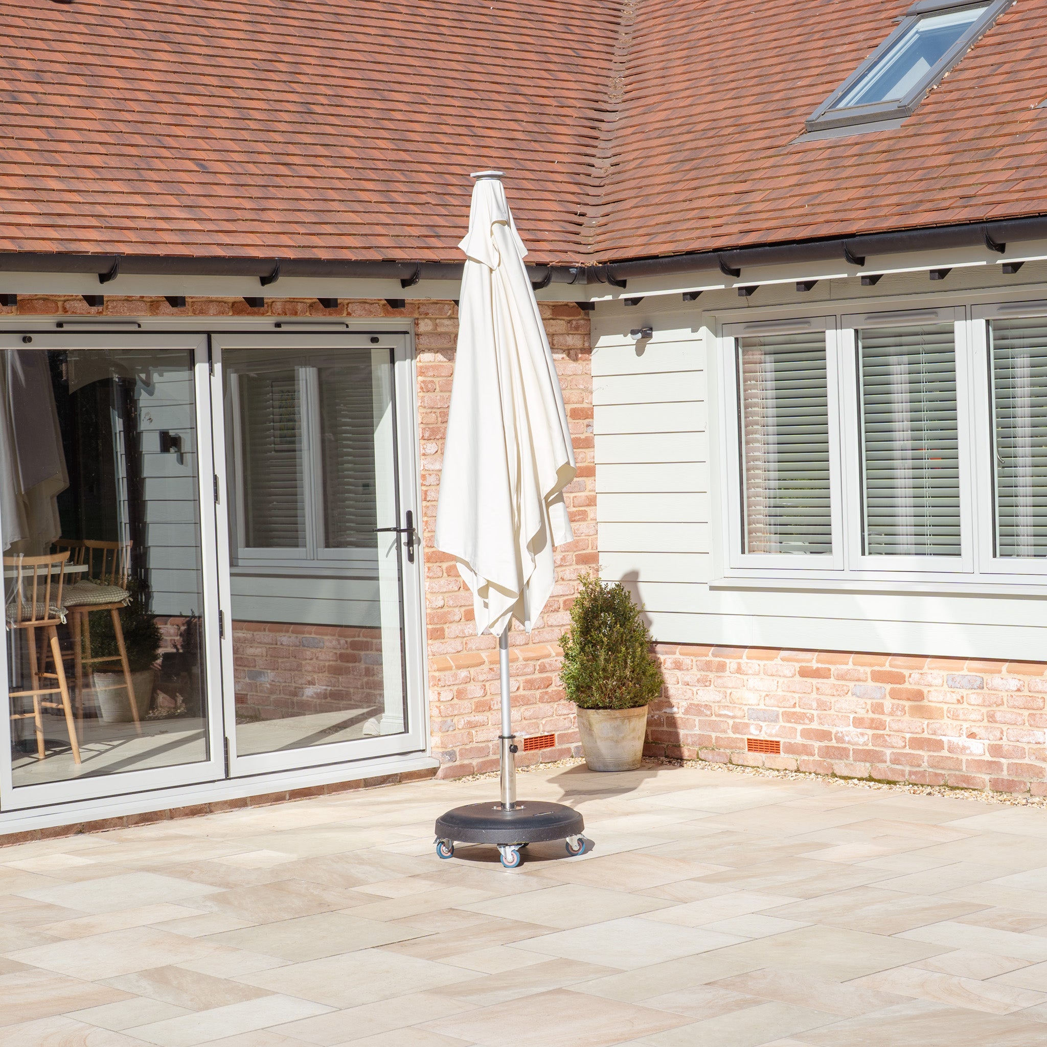 The Zelos 3m x 2m Rectangular Parasol in Cream with LED Lighting and UV protection sits closed on a wheeled stand near a brick house with large glass doors and windows.