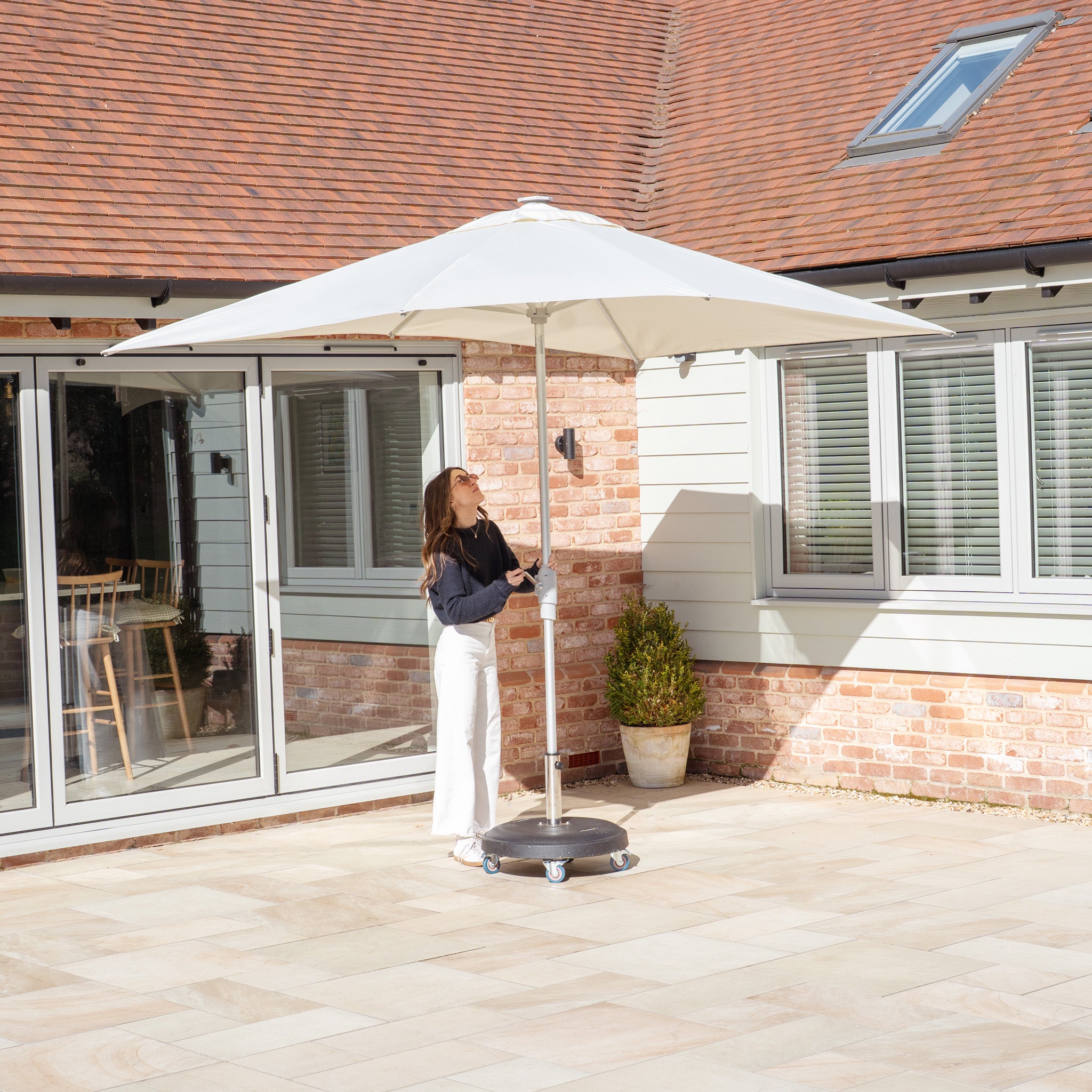 A person adjusts the Zelos 3m x 2m Rectangular Parasol with LED Lighting in Cream on a sunny brick and siding patio near a house with glass doors.