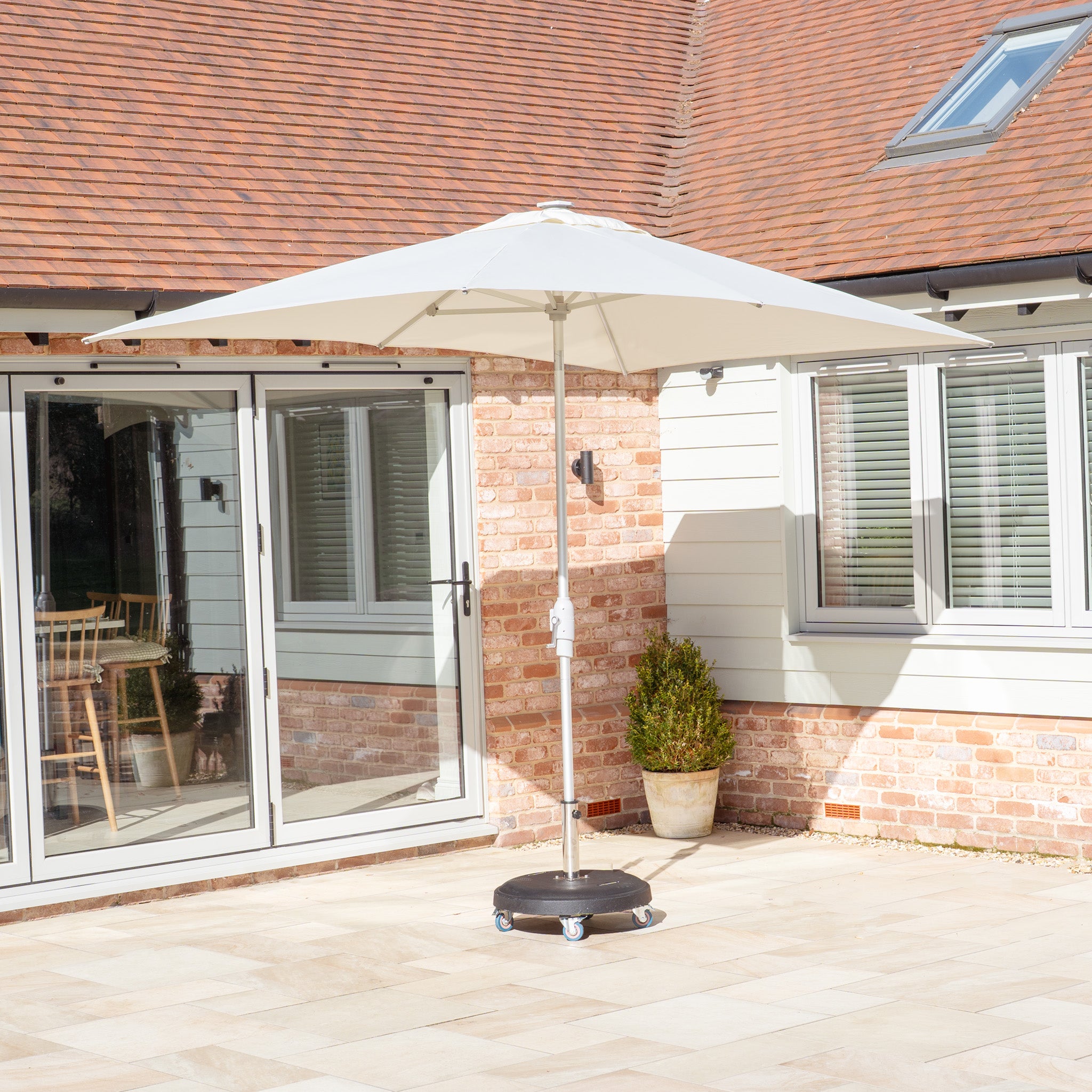 A cozy patio features a Zelos 3m x 2m Rectangular Parasol with LED Lighting in Cream, surrounded by beige brick walls and glass doors. A potted plant rests by the window, soaking in soft natural light under the large umbrella.