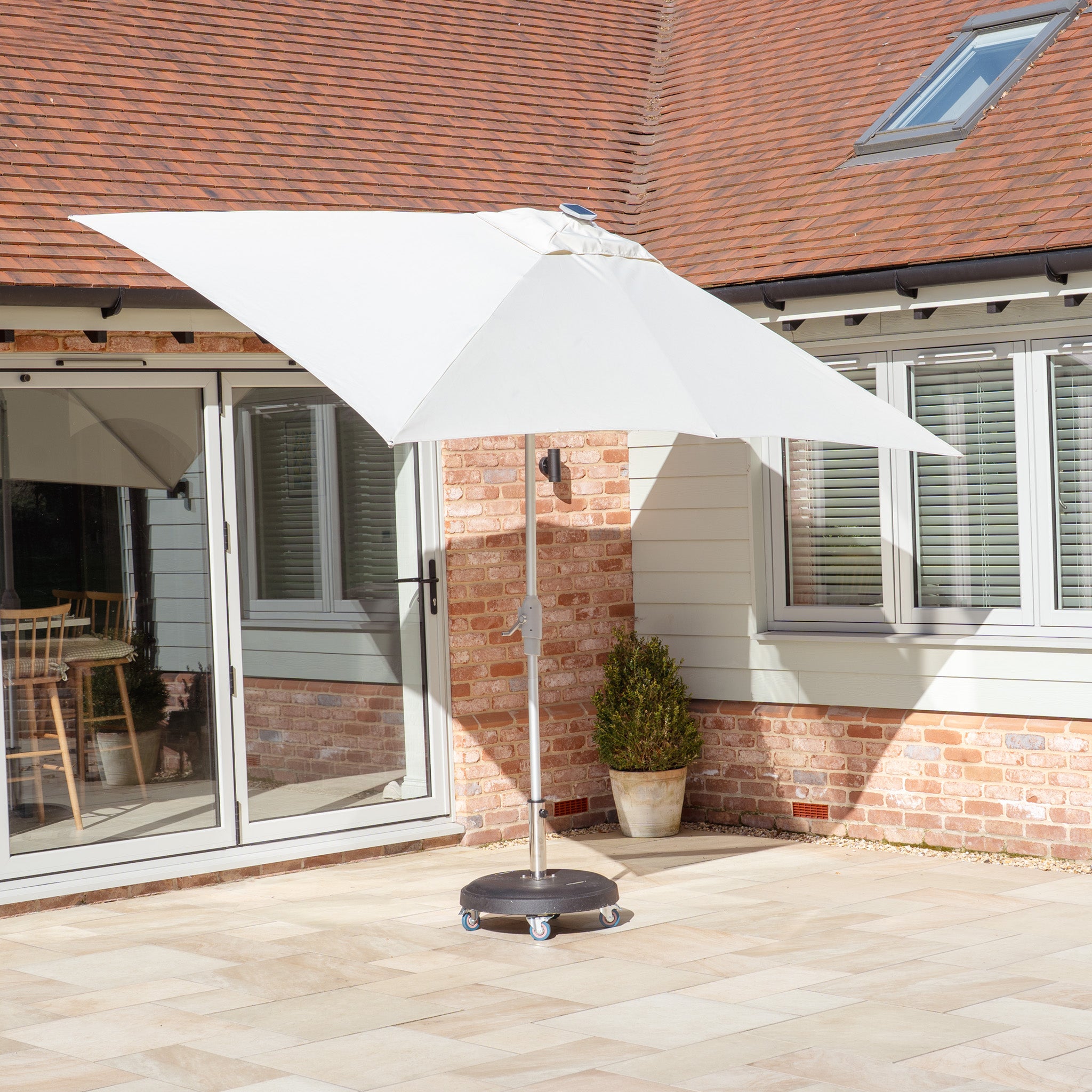 A Zelos 3m x 2m Rectangular Parasol with LED Lighting in Cream adorns the tiled outdoor area near a house with large windows and a brown roof, offering UV protection and evening ambiance.