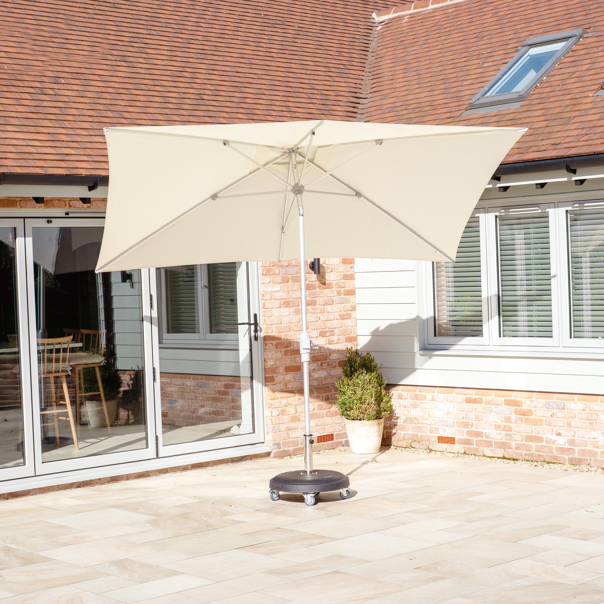 In a sunlit courtyard with brick walls and large windows, the Zelos 3m x 2m Rectangular Parasol with LED Lighting in Cream stands on a base, providing UV protection to create the perfect shaded oasis.