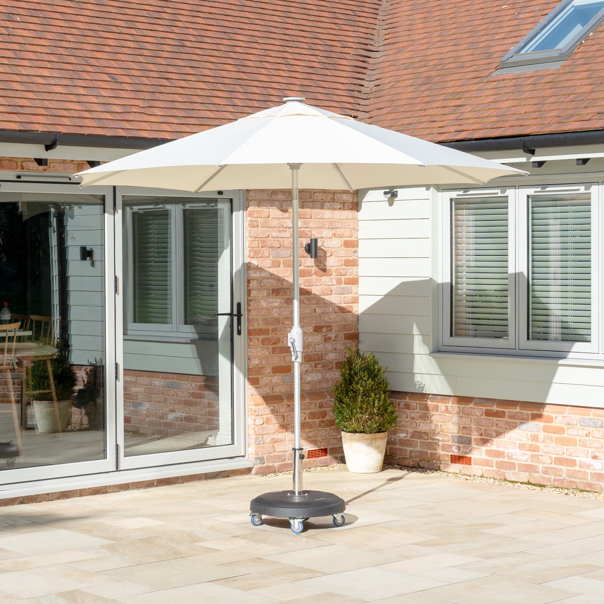 A sunny backyard features a patio with a large cream Zelos 2.5M Round Parasol with LED lighting providing UV protection, complemented by a potted plant and glass doors, enhancing the ambiance for relaxing evenings outdoors.