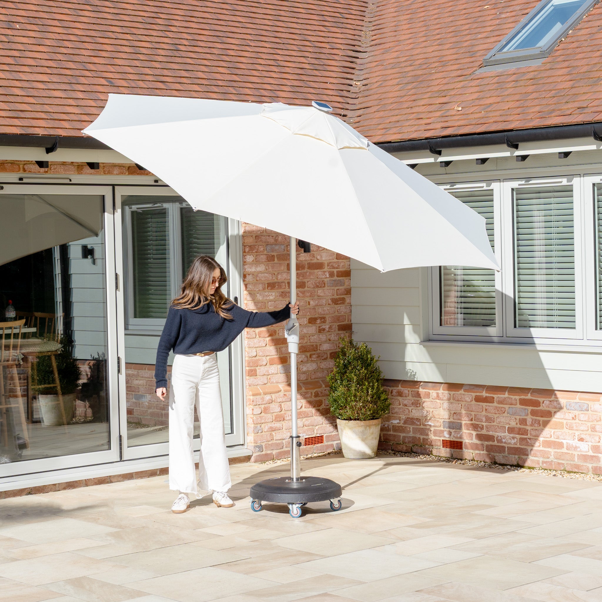 A person adjusts the Zelos 2.5M Round Parasol with LED Lighting in Cream on a sunny terrace, surrounded by brick walls and windows.