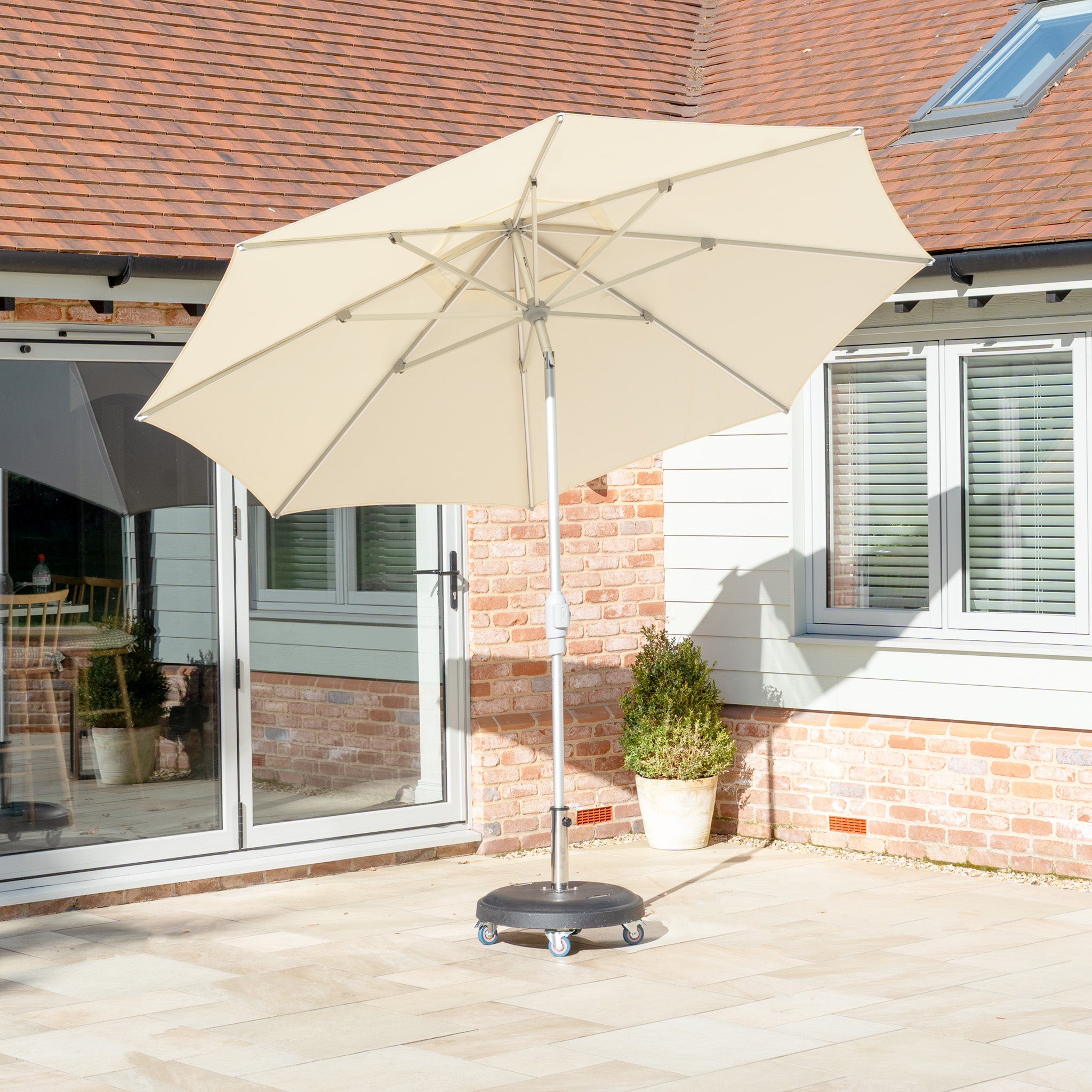 The Zelos 2.5M Round Parasol in cream, featuring LED lighting, offers UV protection. Its wheeled base complements the sunny patio by the house's charming brick and siding exterior, perfect for outdoor living.