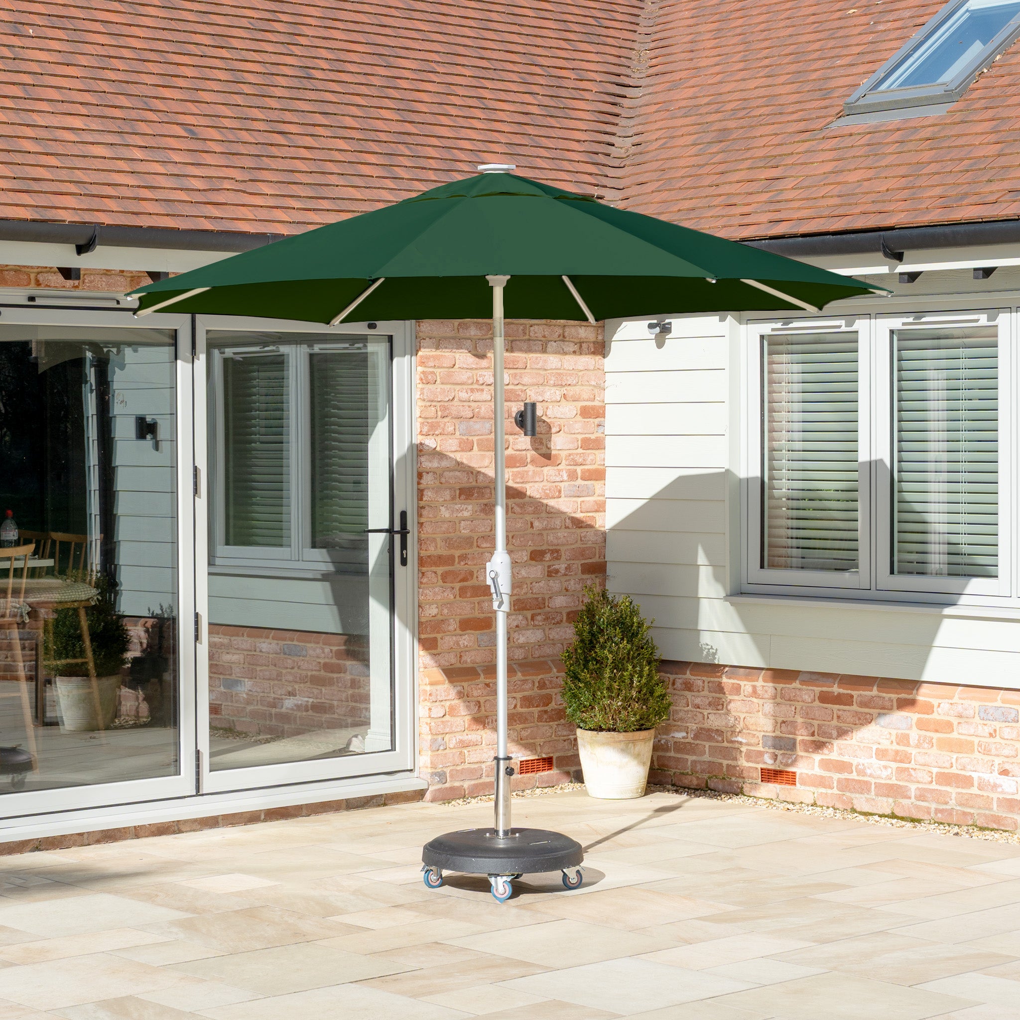 The Zelos 2.5M Round Parasol with LED Lighting in Green offers ideal outdoor shade on the tiled terrace, featuring a sturdy metal stand by the house with brick and light siding.