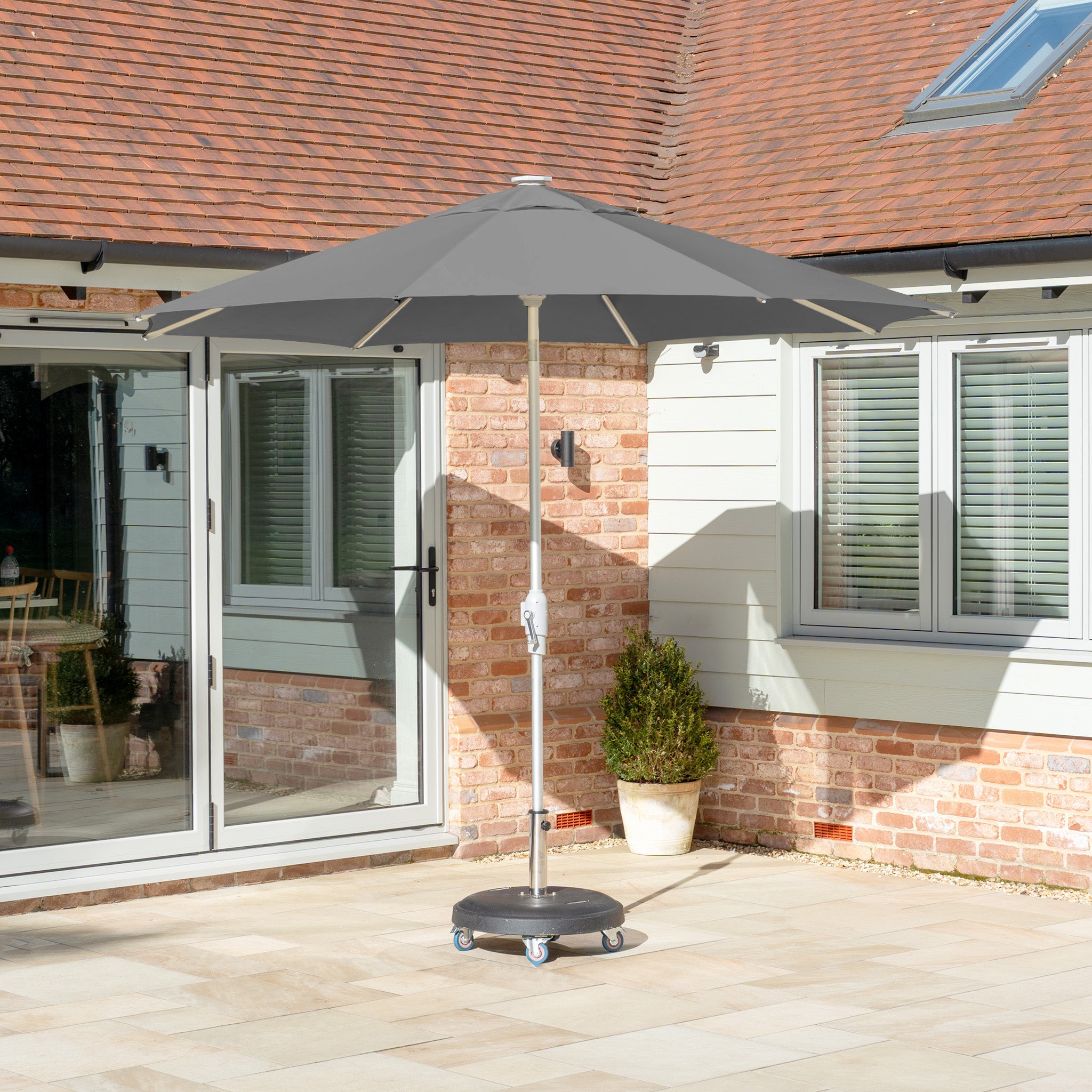 A gray Zelos 2.5m round parasol with LED lighting, featuring an aluminum frame for UV protection, stands on a wheeled base in a sunlit courtyard near a brick and paneled house.
