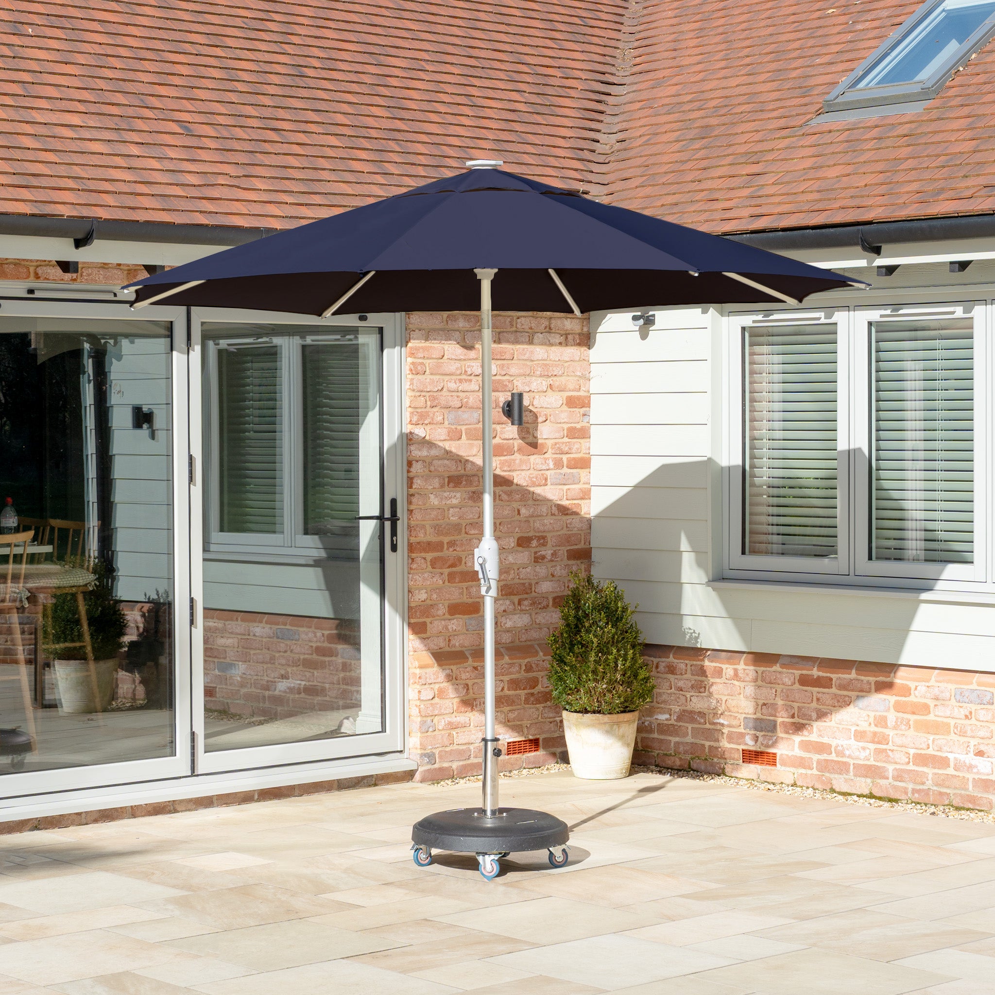 The Zelos 2.5M Round Parasol with LED Lighting in Navy enhances the sunlit stone patio, beautifully complementing the house's glass doors and brick walls with its fade-resistant fabric for lasting outdoor charm.