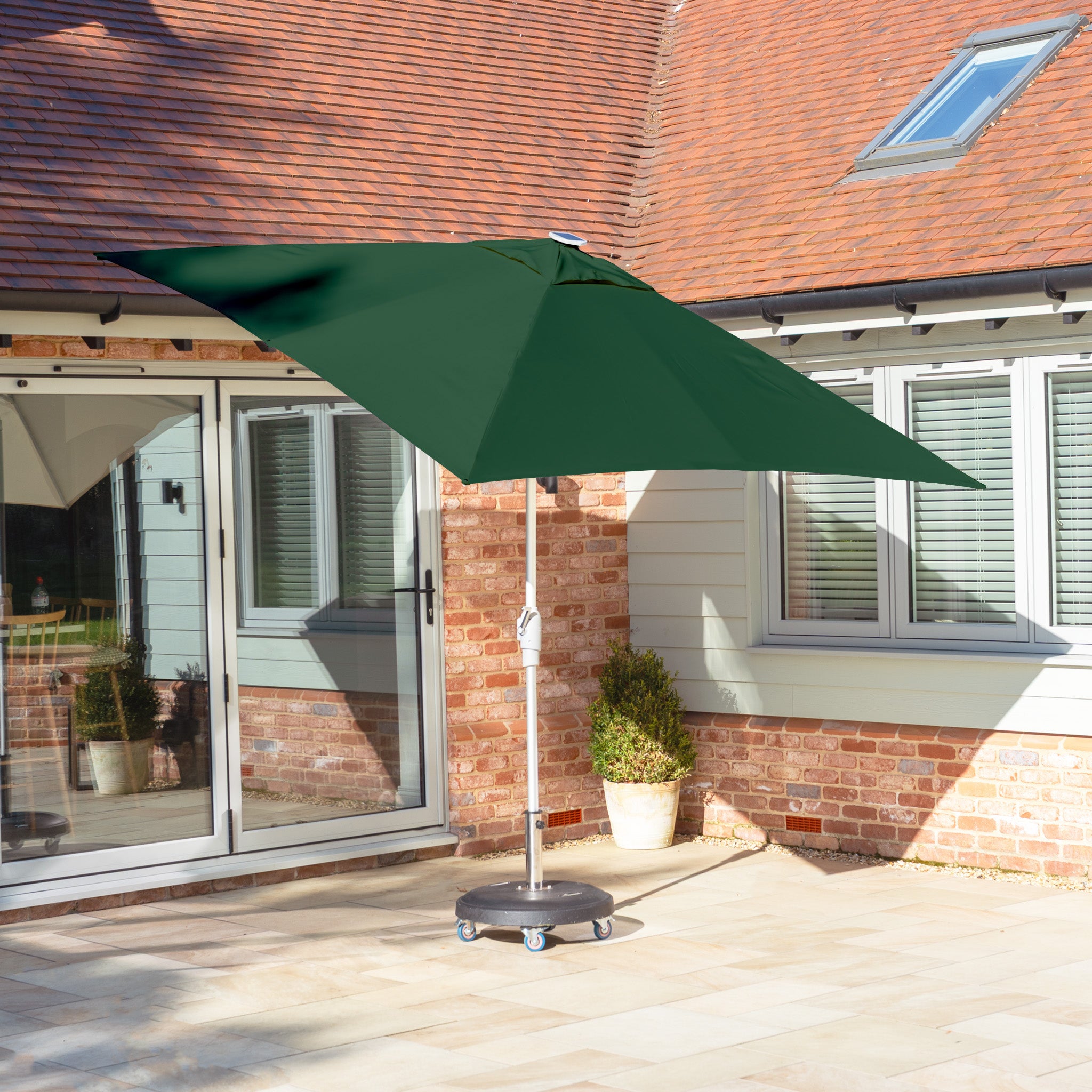 A Zelos 2.5m Square Parasol with LED Lighting in Green is placed on a sunlit stone terrace near a house with brick and siding walls, illuminating the outdoor space.
