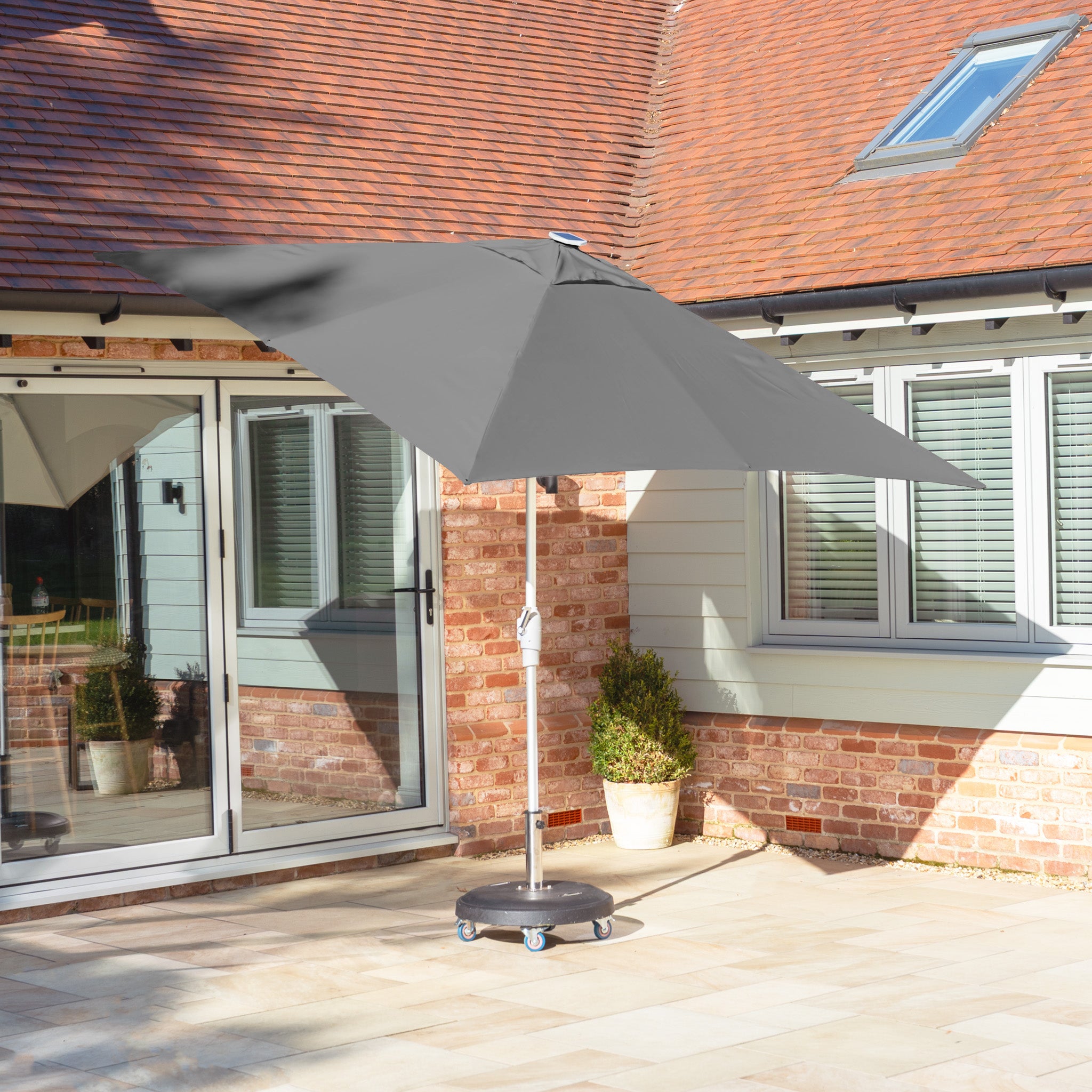 The Zelos 2.5m Square Parasol with LED Lighting in Grey adds elegance to the stone terrace, prominently situated beside a brick wall and sliding glass doors.