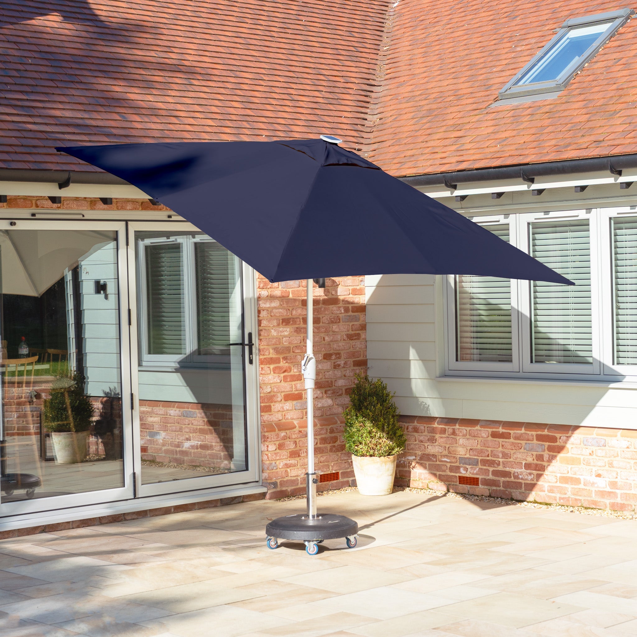The patio is adorned with stone tiles and a brick wall, accentuated by sliding glass doors. The Zelos 2.5m Square Parasol with LED Lighting in Navy offers shade and adds a touch of style, enhancing the area's appeal.