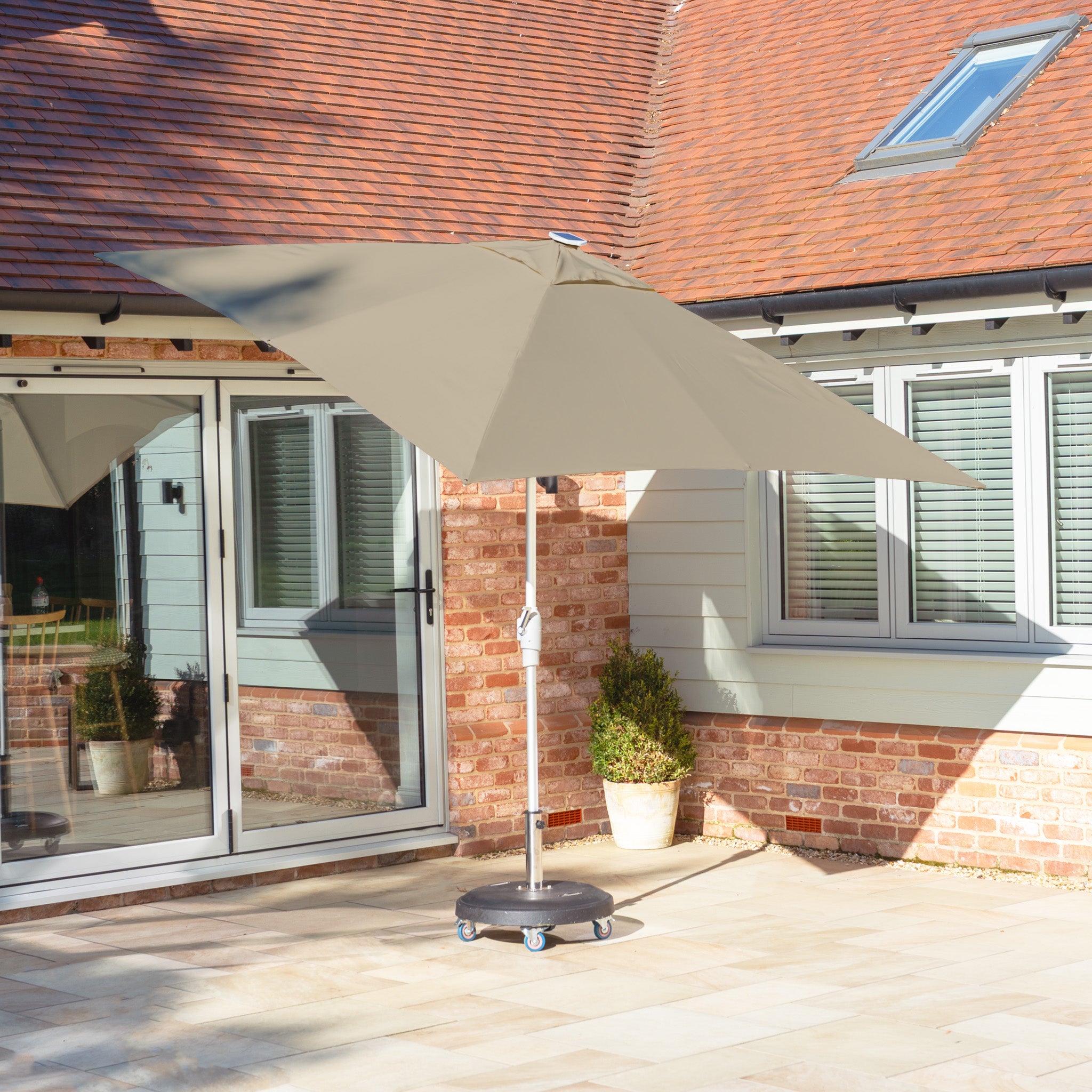 Patio featuring a Zelos 2.5m Square Parasol with LED Lighting in Taupe, sliding glass doors, a brick wall, and potted plants by a house with a tiled roof.