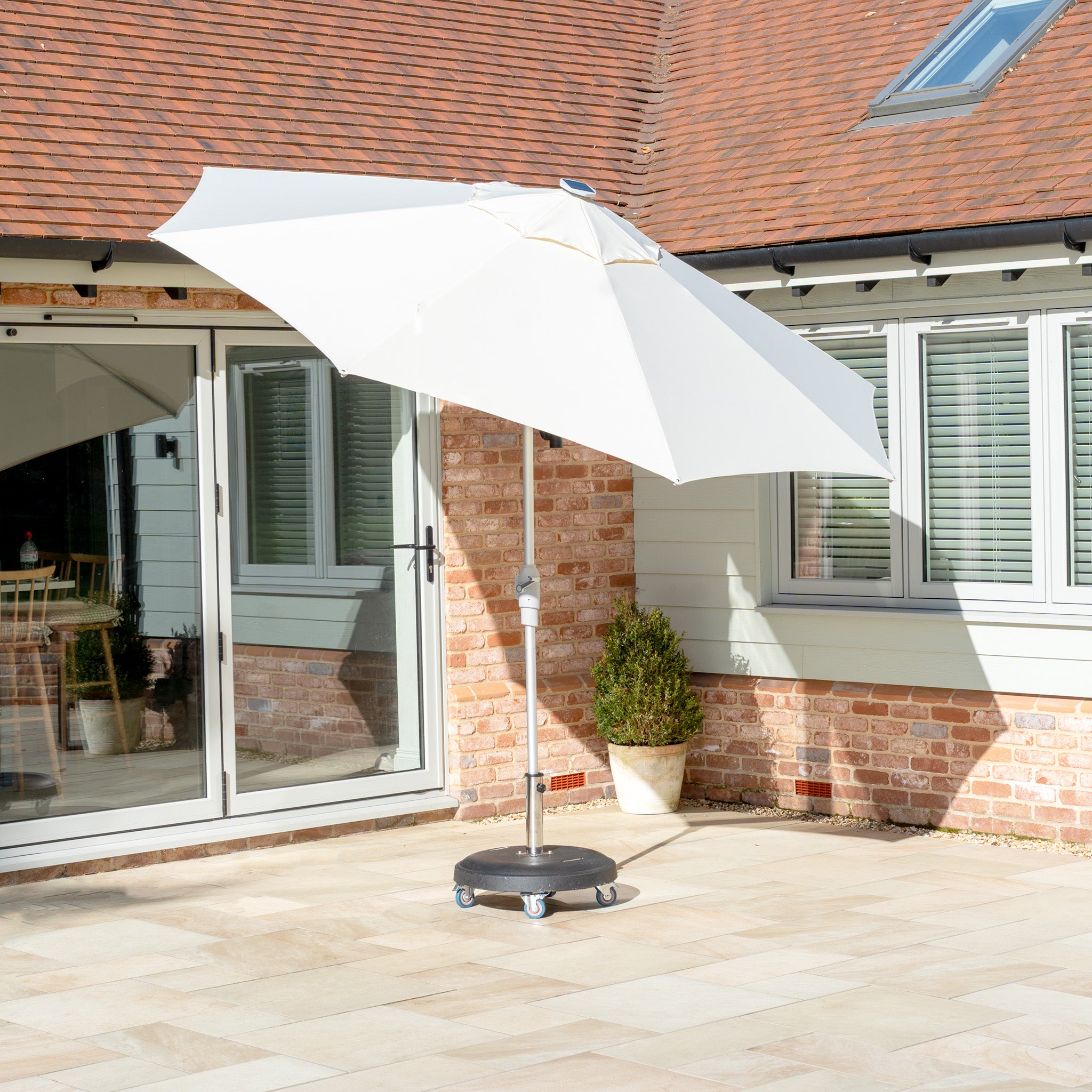 A Zelos 3m Round Parasol in cream with LED lighting and UV protection is set on a sunny paved patio near a house with brick and glass walls, featuring a stand with wheels.