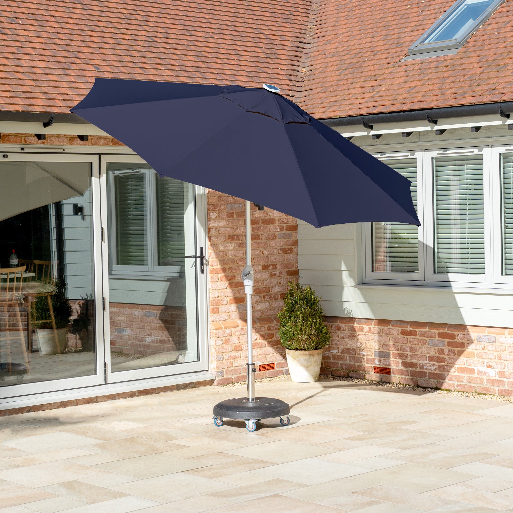 A navy Zelos 3m round parasol with LED lighting and UV protection sits on a wheeled base on a stone patio near a brick house with glass doors.