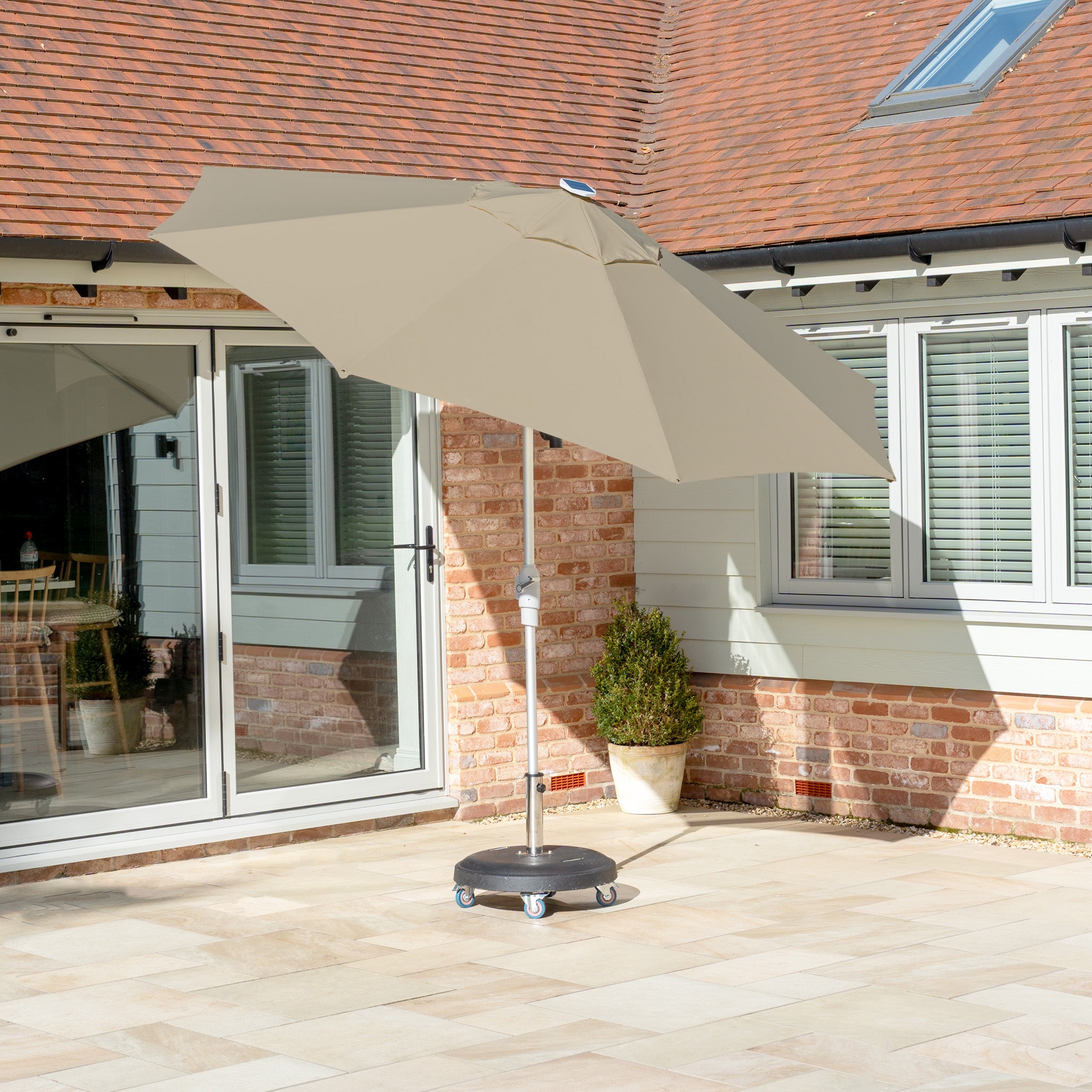 The Zelos 3m round parasol with LED lighting in taupe graces the patio, offering shade and UV protection. Brick walls and sliding glass doors frame this serene corner, creating an inviting backdrop for leisurely afternoons.