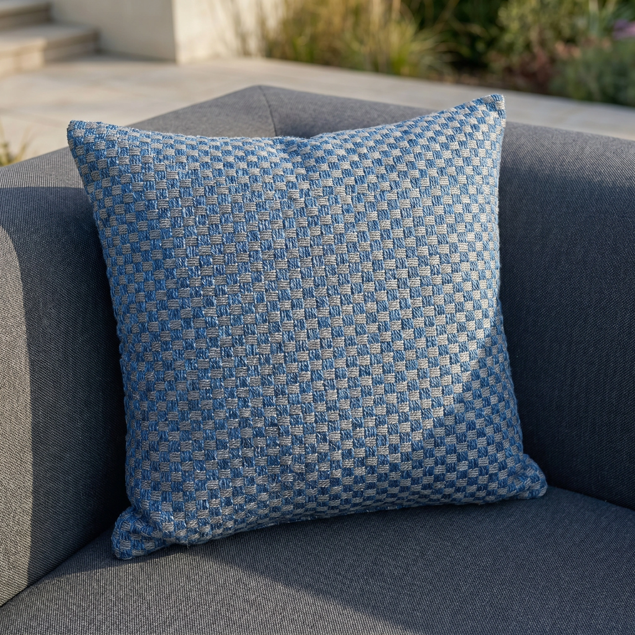 The Agora Senda Blue Medium Scatter Cushion (45cm x 45cm) adds a blue and gray checkered touch to a gray outdoor armchair in a sunlit setting.