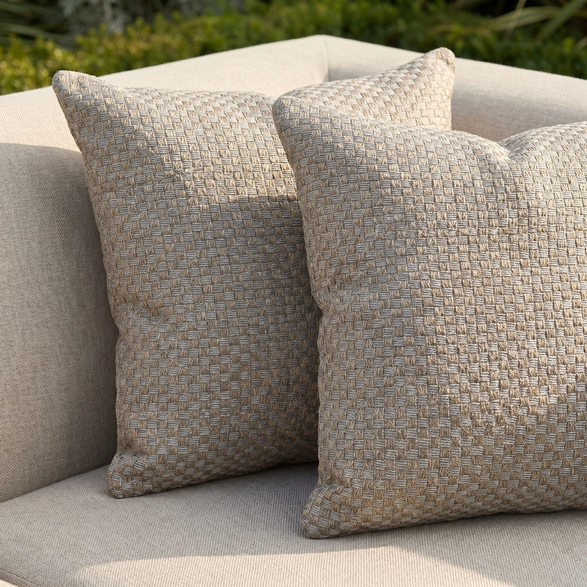Two Agora Senda Taupe Medium Scatter Cushions (45cm x 45cm), crafted from solution-dyed acrylic, sit on a light outdoor sofa with lush greenery in the background.