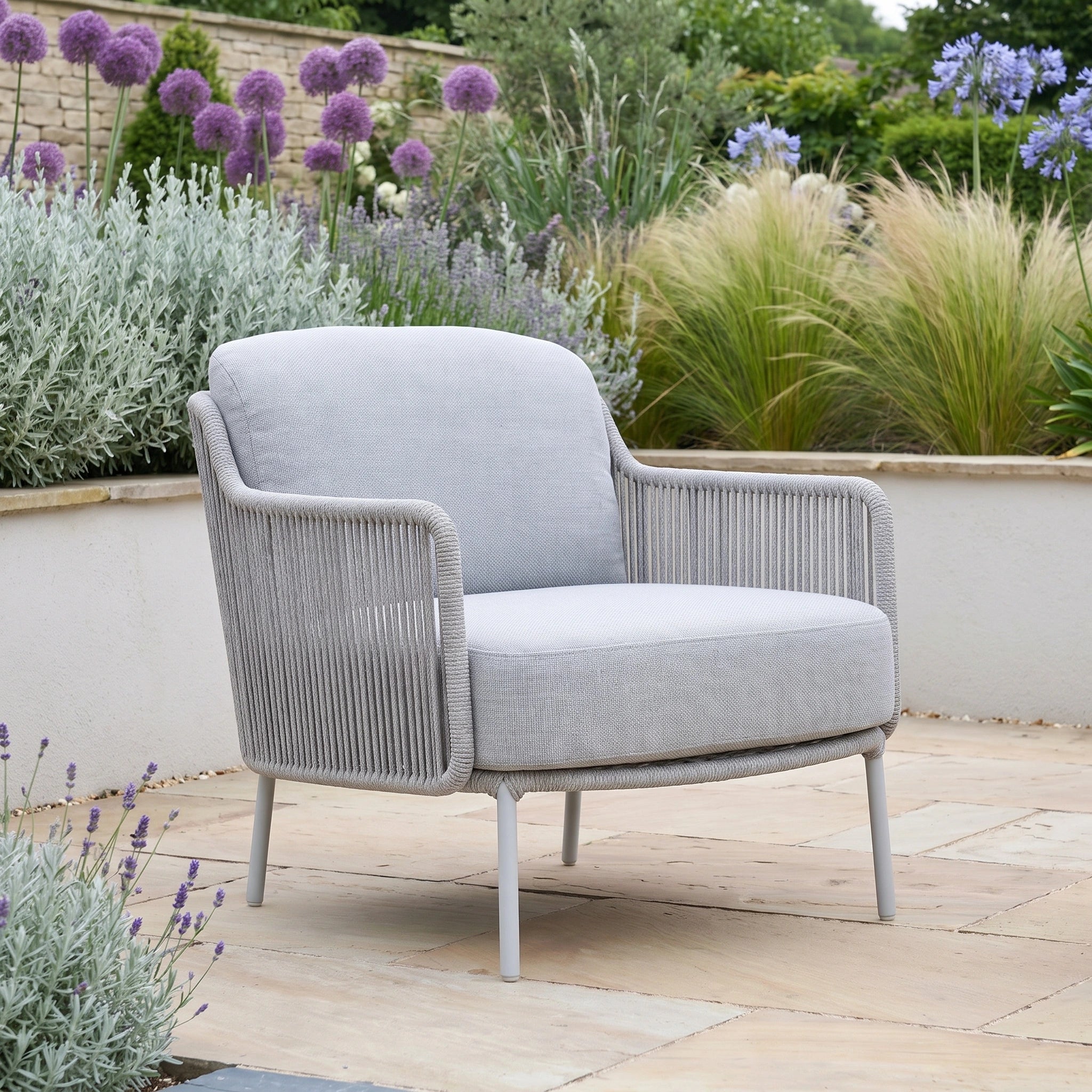 A Bernini 2 Seat Sofa Set with Teak Tables and Footstool in Light Grey sits on a stone patio, surrounded by flowering garden plants and greenery, showcasing the elegance of handmade outdoor furniture.