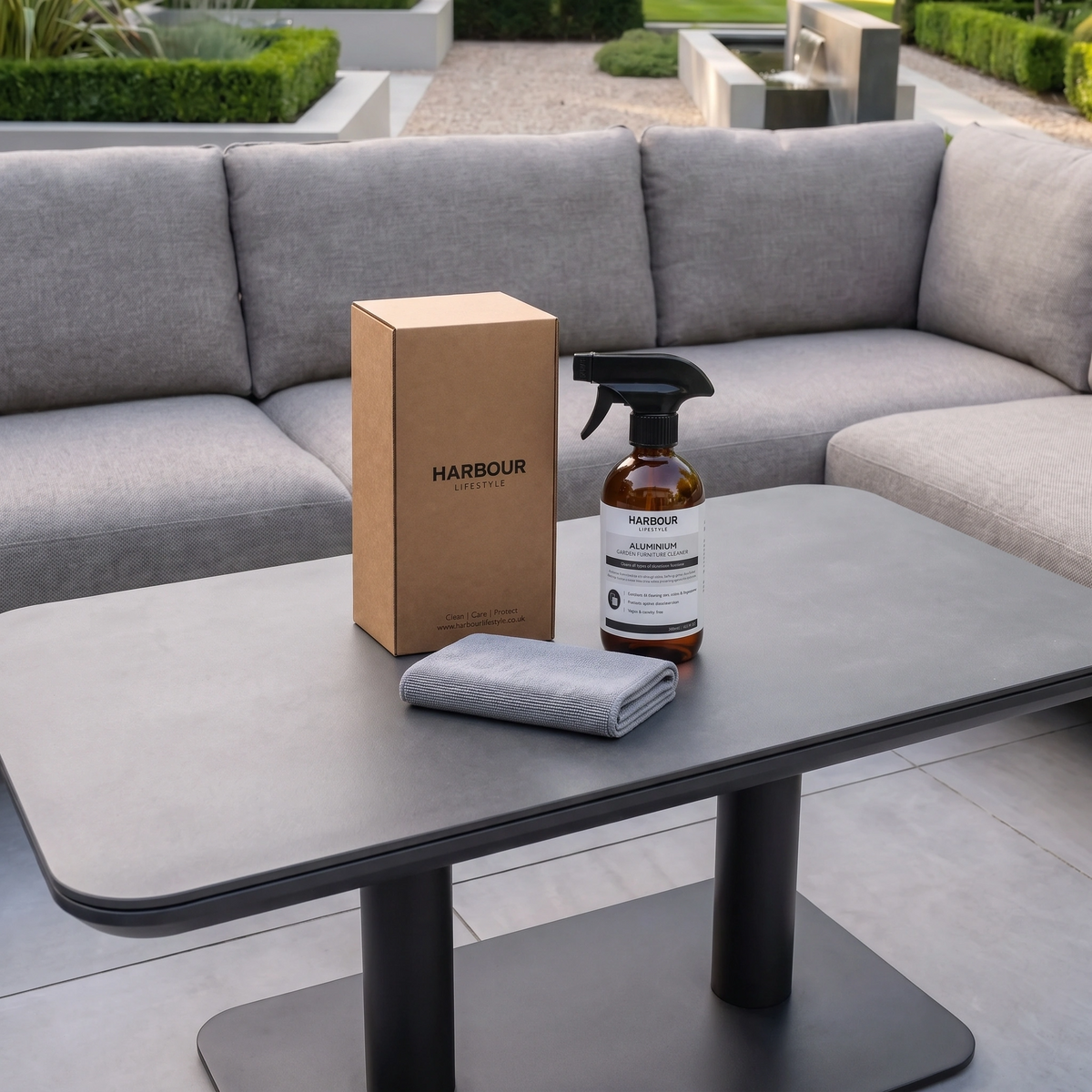 A spray bottle, cloth, and box of Outdoor Aluminium Furniture Cleaner rest on a dark table in front of a gray outdoor sofa.