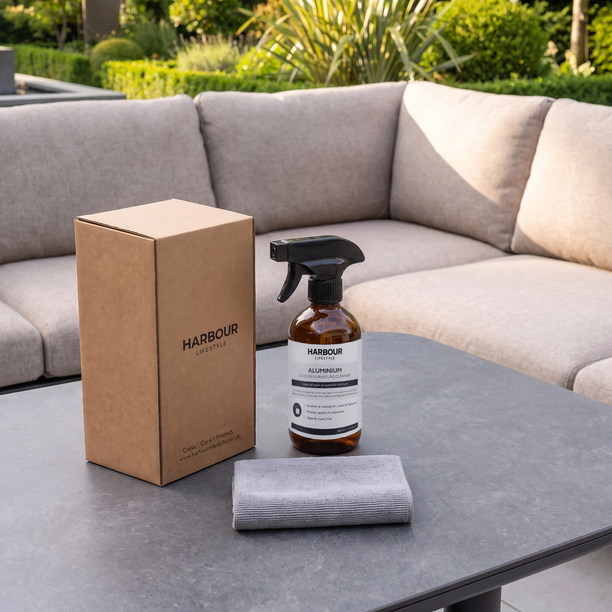 A box, spray bottle of Outdoor Aluminium Furniture Cleaner, and a gray cloth sit on a gray outdoor table with a sofa and greenery in the background.