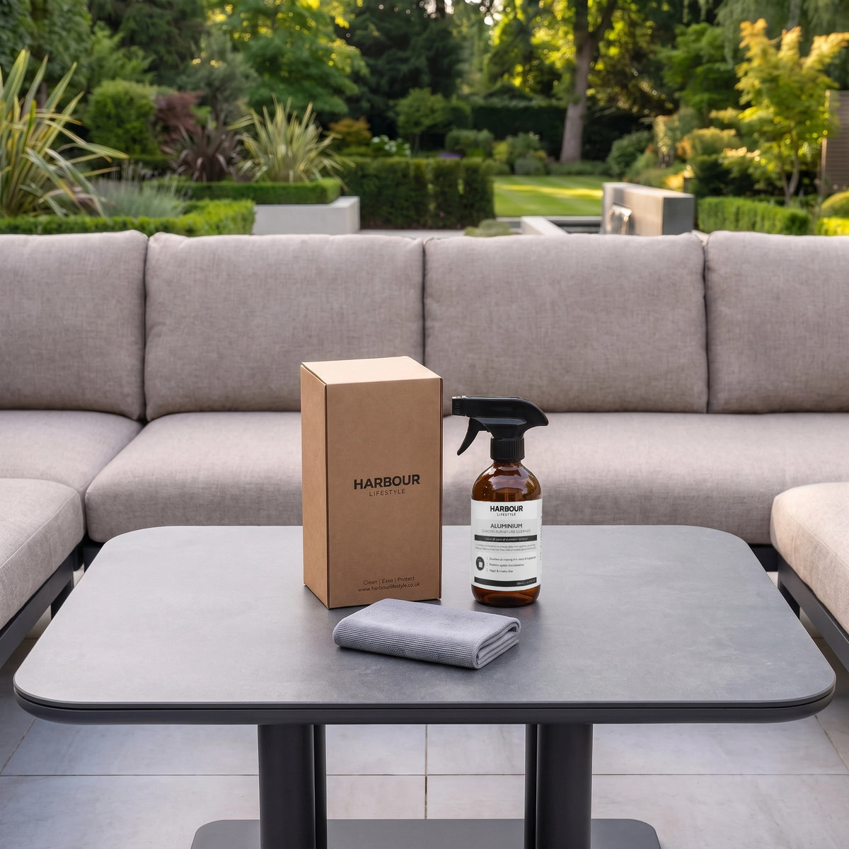 A box, spray bottle, and cloth on a table in front of an outdoor sofa set in a lush green garden—ready for use with your favourite Outdoor Aluminium Furniture Cleaner.