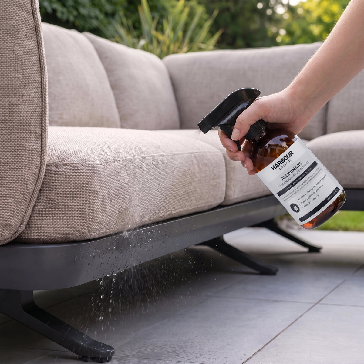 Spraying Outdoor Aluminium Furniture Cleaner onto the base of an outdoor sofa on a tiled patio, with greenery in the background.