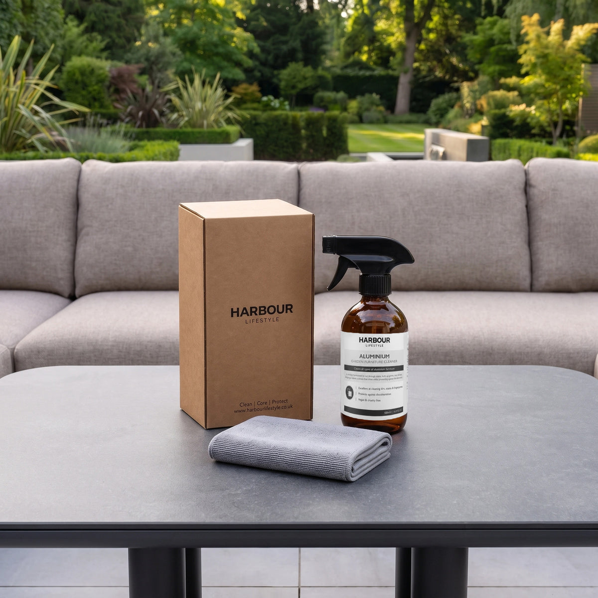 A spray bottle, box, and gray cloth rest on a table outdoors with a sofa and greenery in the background—ideal for using your Outdoor Aluminium Furniture Cleaner to keep your outdoor space looking fresh.