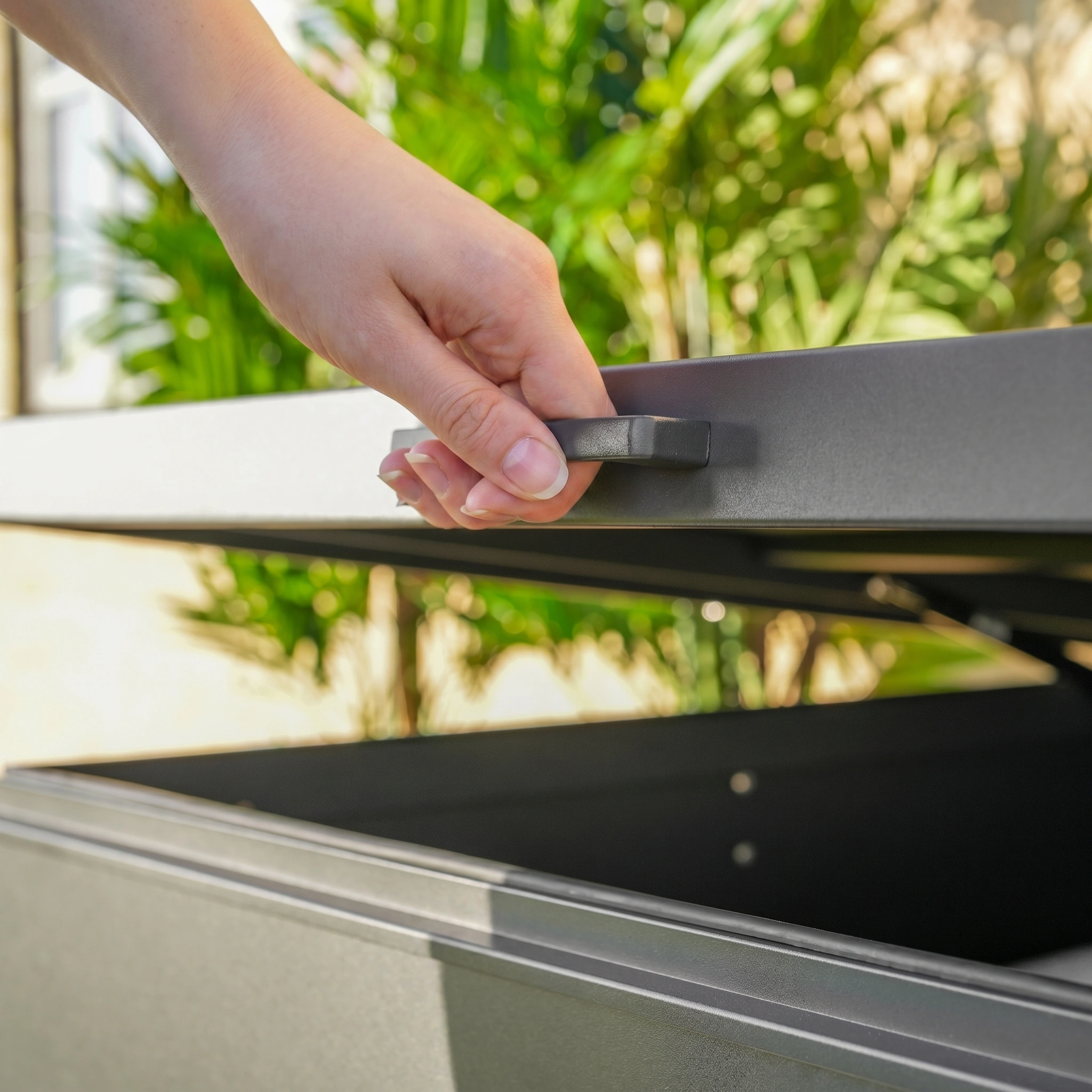 A hand lifts the lid of the Large Aluminium Storage Box in Grey with green plants behind, highlighting this stylish and practical waterproof garden box for backyard essentials.