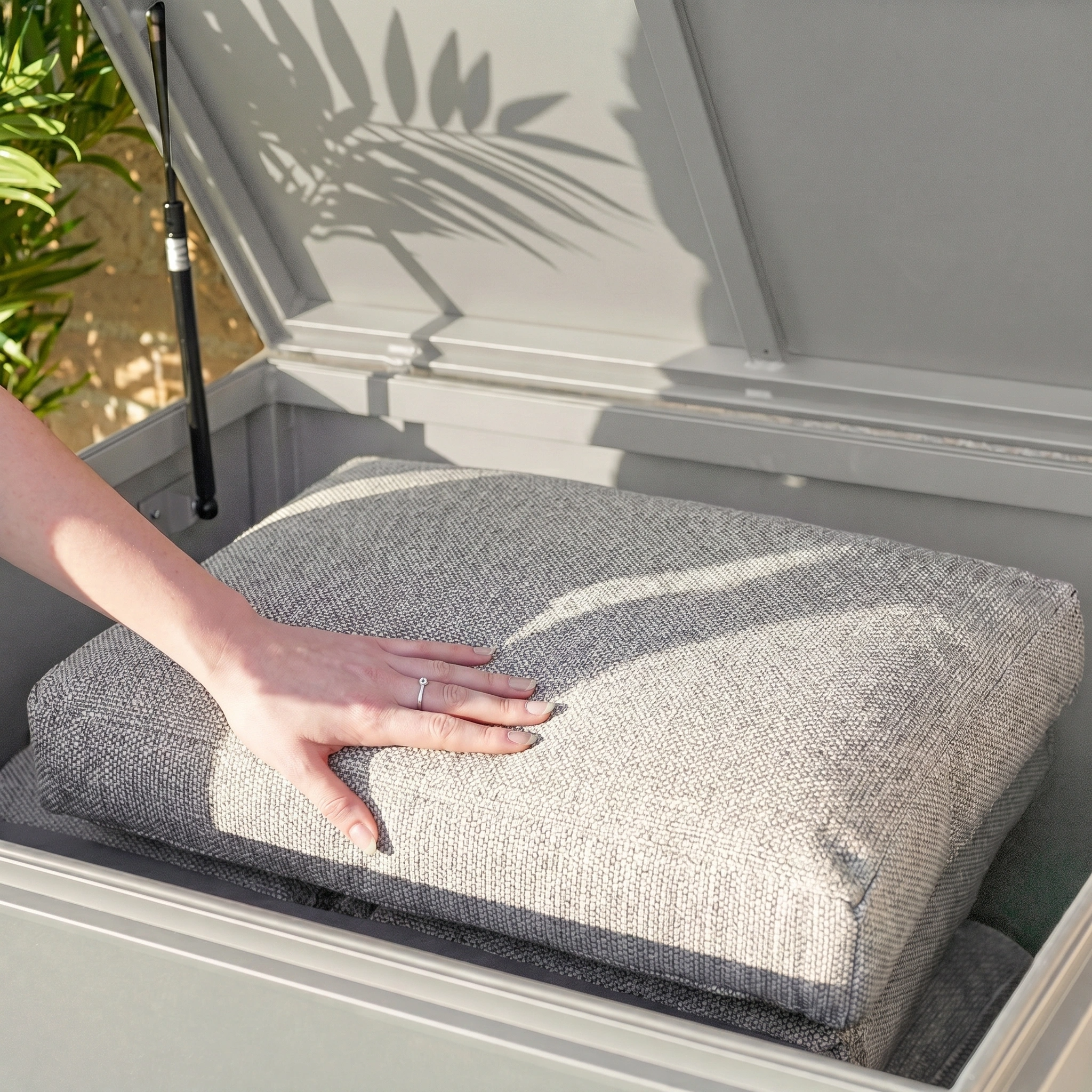 A hand places a cushion inside the Large Aluminium Storage Box in Latte, with sunlight and plant shadows highlighting its spacious design.