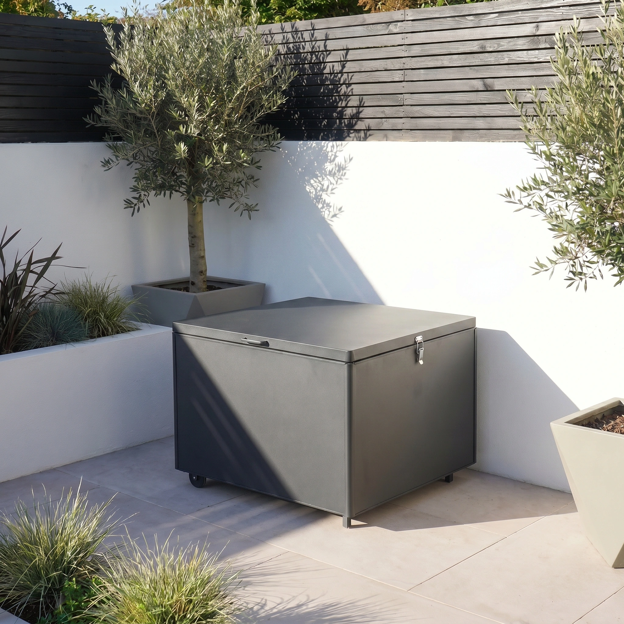 The Small Aluminium Storage Box in Grey, featuring wheels, provides stylish and practical waterproof garden storage on a tiled patio beside potted plants and an olive tree.