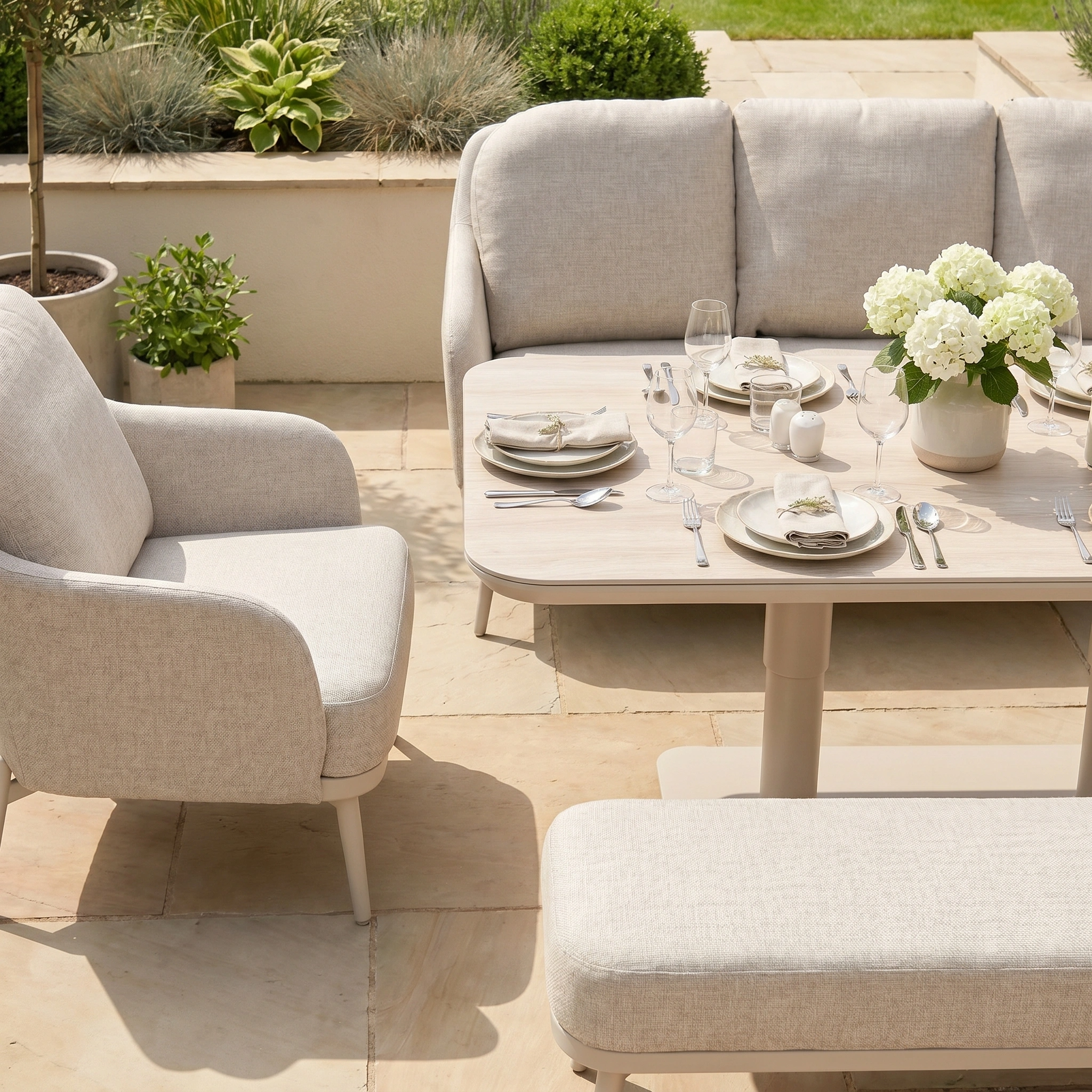 Stylish outdoor dining set featuring the Alva 3 Seat Sofa Dining Set with Rising Table in Fawn, accented with light cushions on a stone patio amidst lush greenery—ideal for garden furniture lovers.