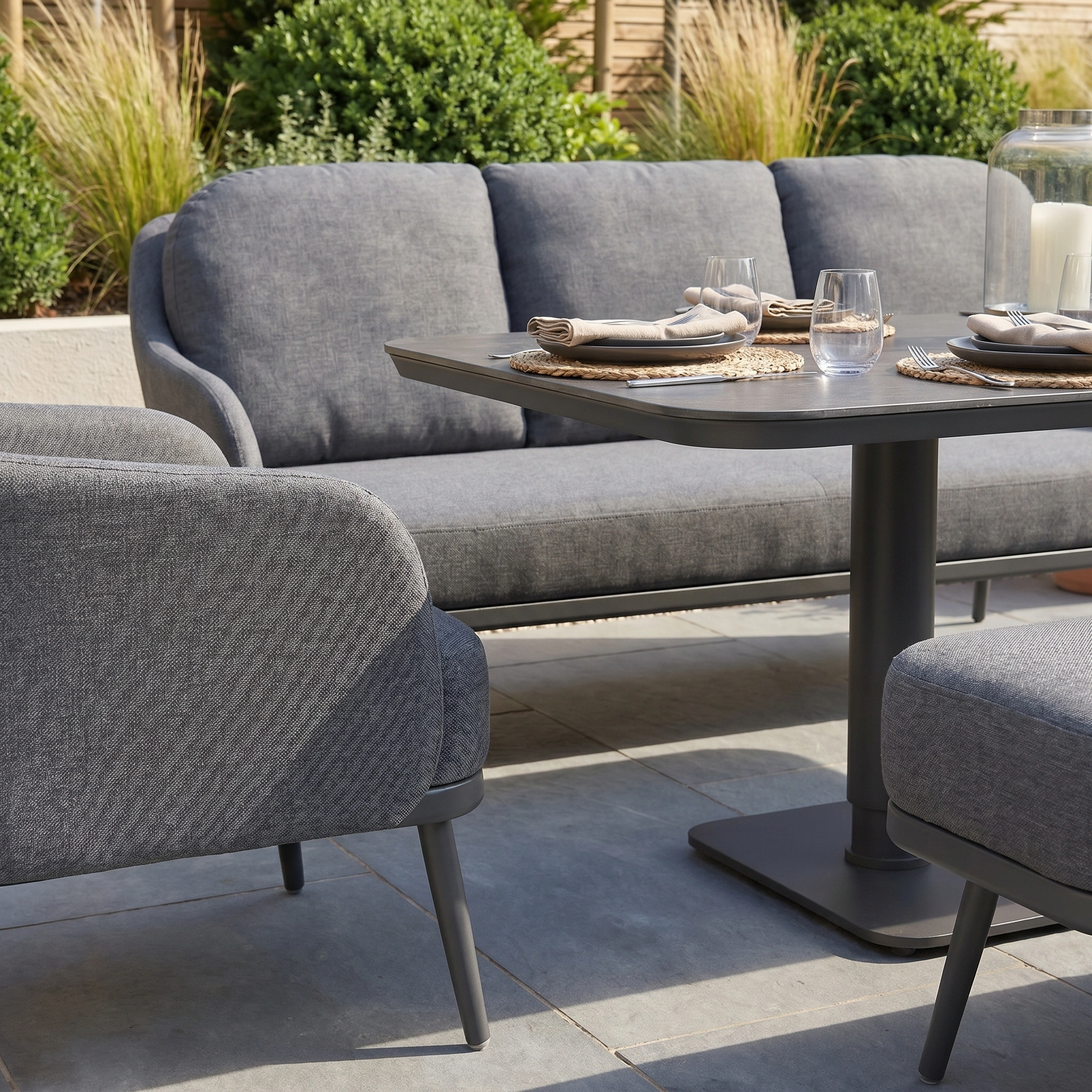 The Alva 3 Seat Sofa Dining Set with Rising Table in Grey offers modern outdoor comfort with cushioned seating and a stylish, adjustable dining table, perfect for relaxing or entertaining amid lush greenery.