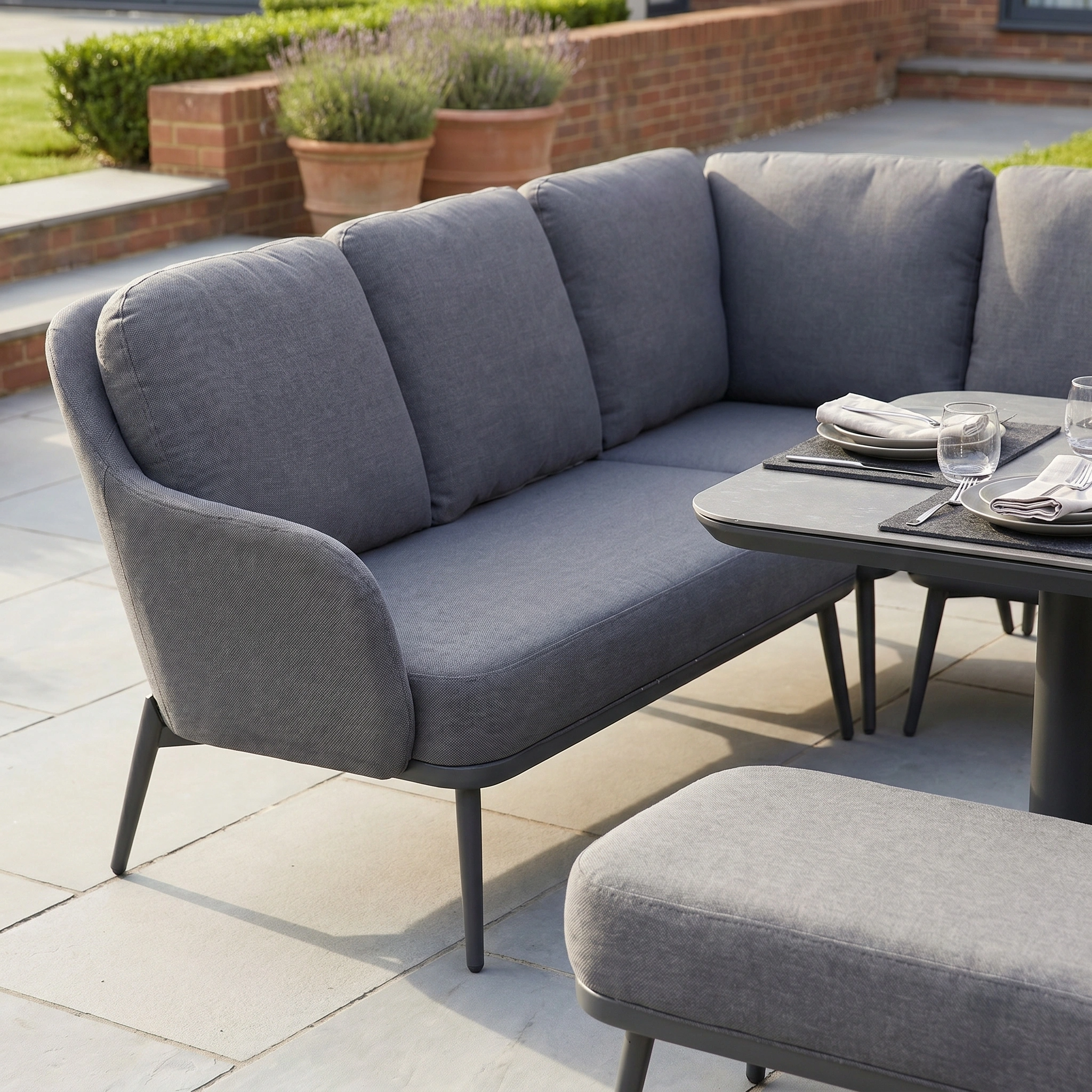 The Alva Corner Dining Set with Rising Table and Armchair in Grey features comfy cushions on a stone patio, surrounded by garden furniture and lush plants.