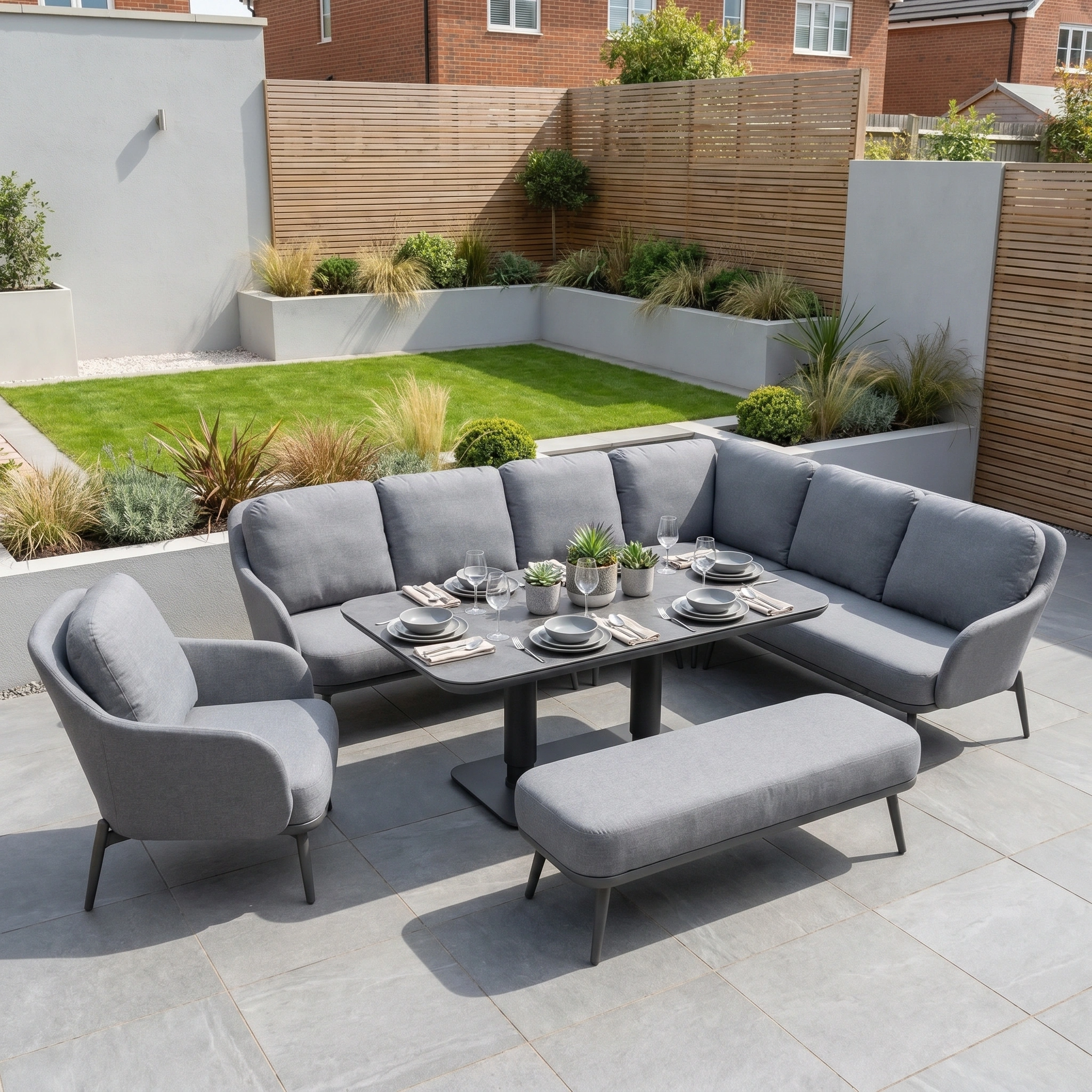 The Alva Corner Dining Set with Rising Table and Armchair in Grey is shown on a tiled terrace, surrounded by a landscaped garden.
