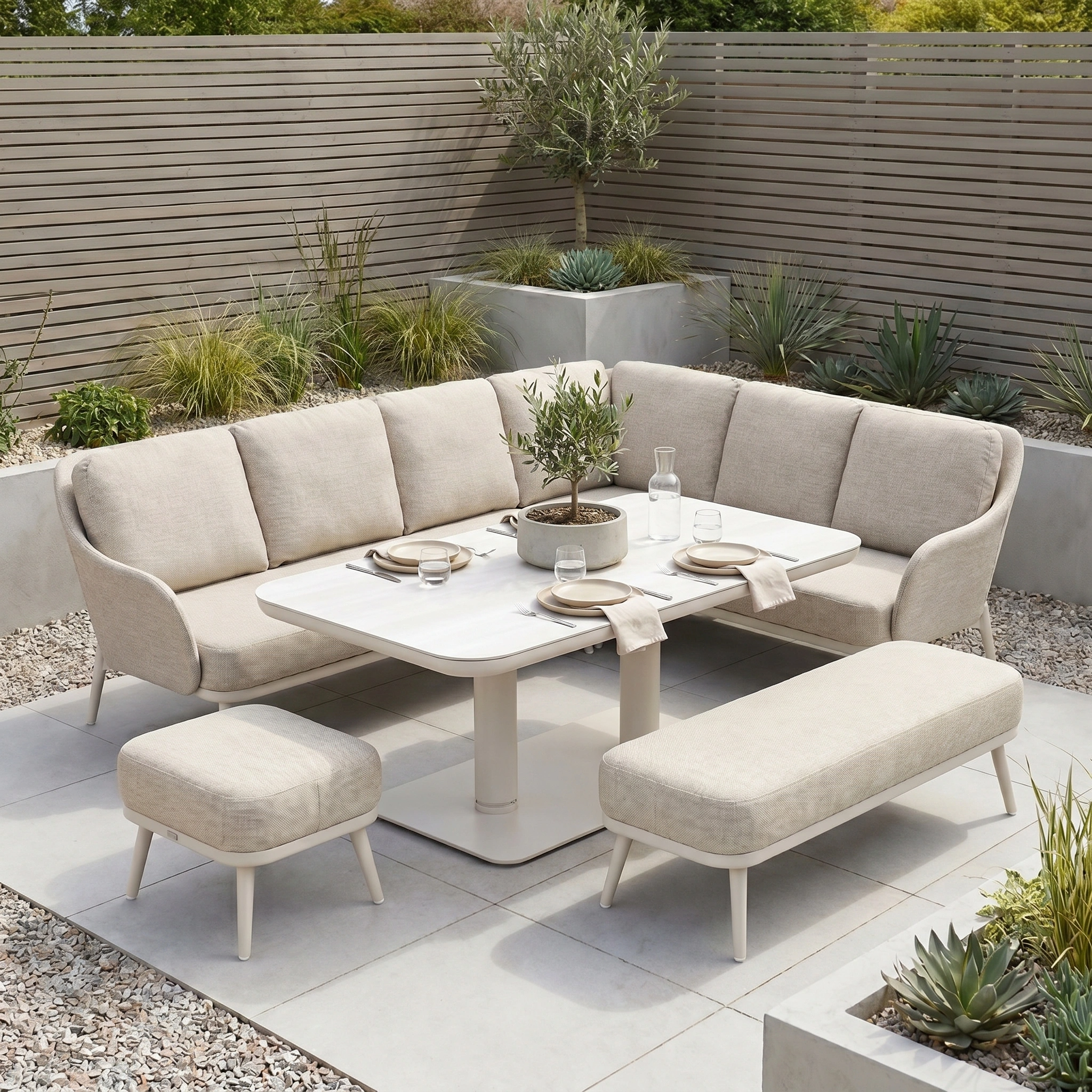 The Alva Corner Dining Set with Rising Table in Fawn features benches and is displayed on a tiled area with plants, surrounded by gravel and greenery.