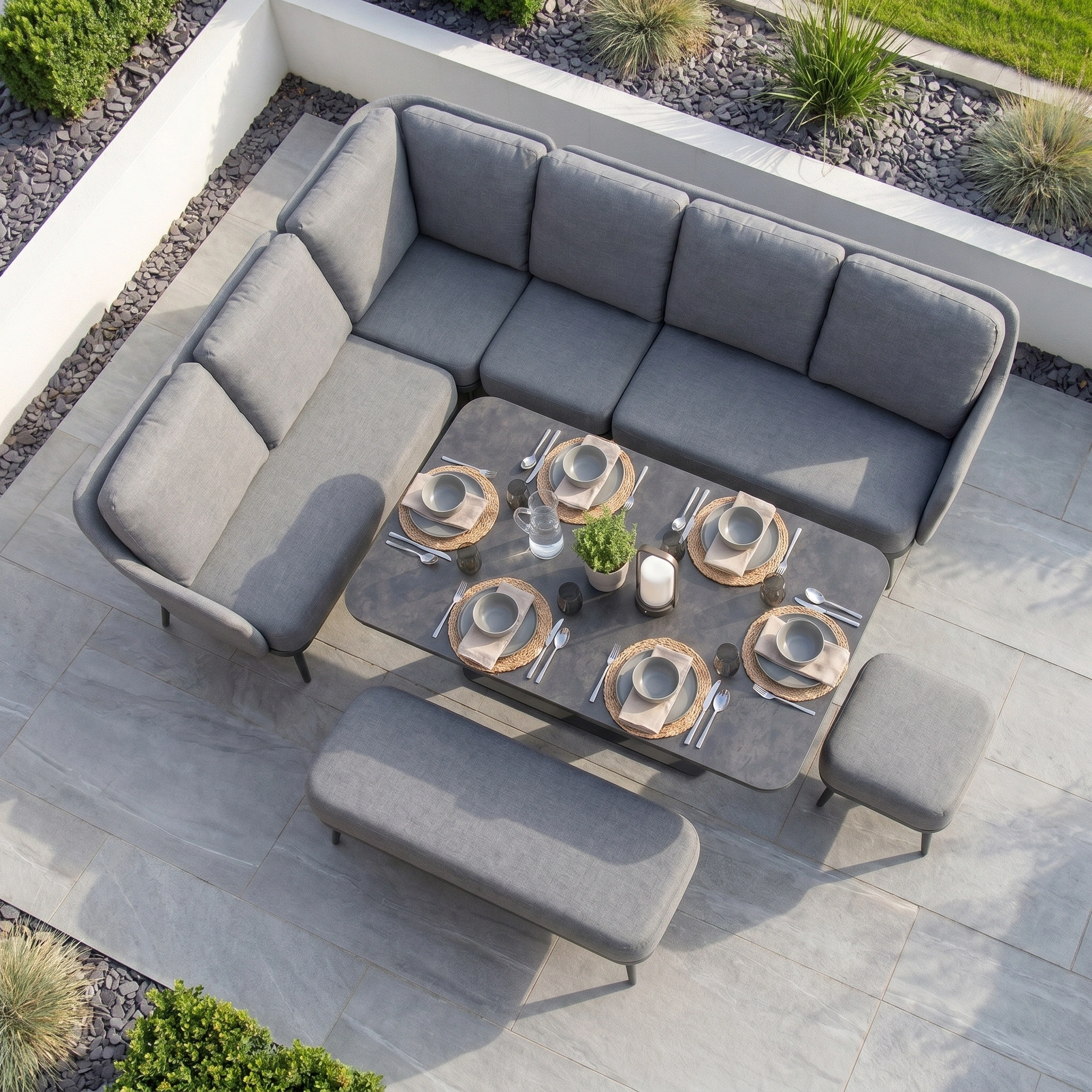 The Alva Corner Dining Set with Rising Table in Grey seats six and offers stylish outdoor comfort, making it perfect garden furniture for a stone patio surrounded by landscaped plants.