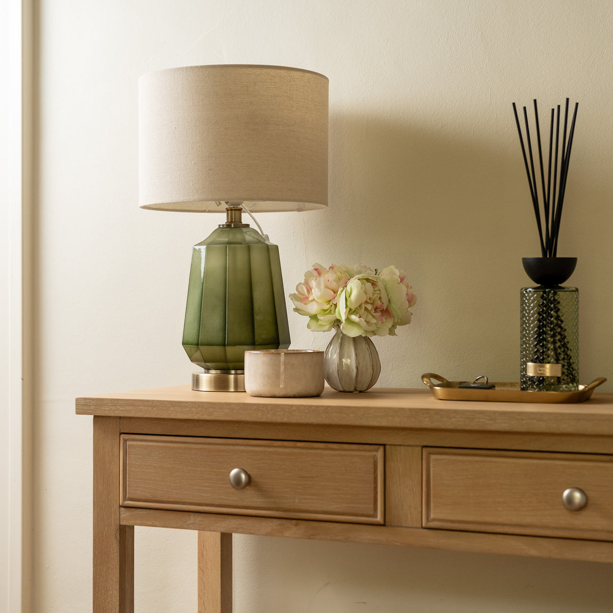 The Amberley 2 Drawers Console Table in Light Oak serves as a stylish entryway accent, topped with a green lamp, a small vase of flowers, a candle, tray, and reed diffuser to create an inviting display. This elegant console table provides both decorative charm and practical storage for your home with its two spacious drawers.